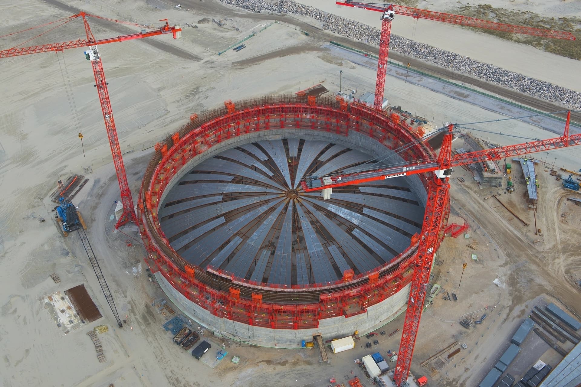 Vue aérienne d'un vaste chantier de construction, montrant une grande structure circulaire en béton avec un toit métallique rayonnant en cours d'installation. Plusieurs grues à tour rouges imposantes sont positionnées autour de l'ouvrage sur un terrain sableux.