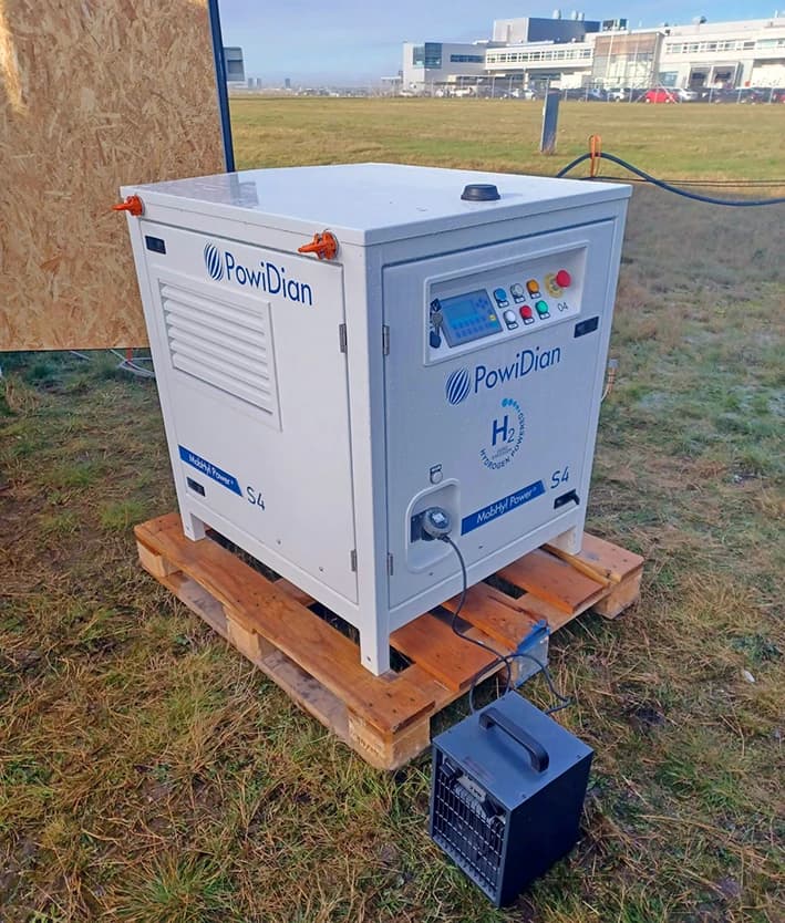 Un générateur de puissance à hydrogène blanc PowiDian MobHyl Power S4 est posé sur une palette en bois sur de l'herbe clairsemée. Un petit appareil gris est connecté au générateur, avec un fond d'aéroport et un ciel brumeux à l'arrière-plan.