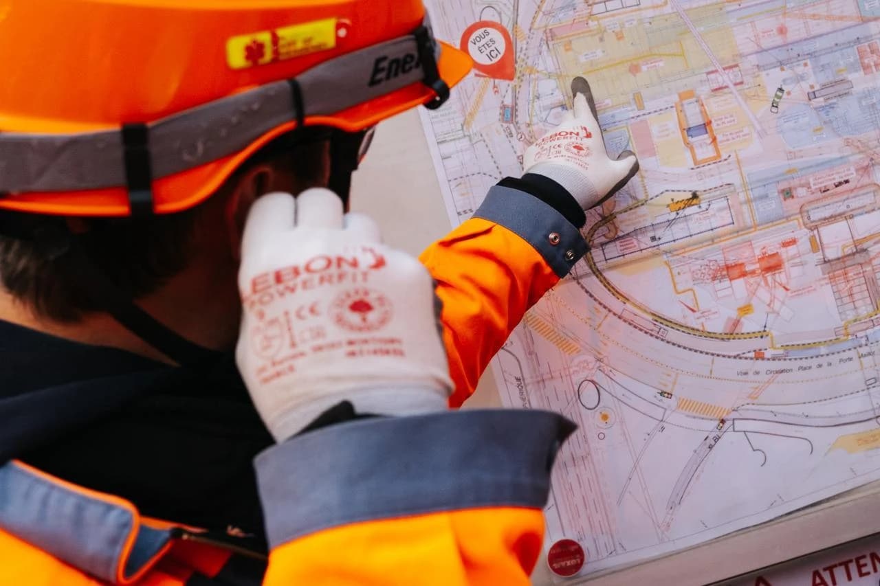 Un travailleur portant un casque et une veste de sécurité orange ainsi que des gants blancs examine attentivement un plan détaillé, pointant du doigt la zone indiquée par un marqueur "VOUS ÊTES ICI".