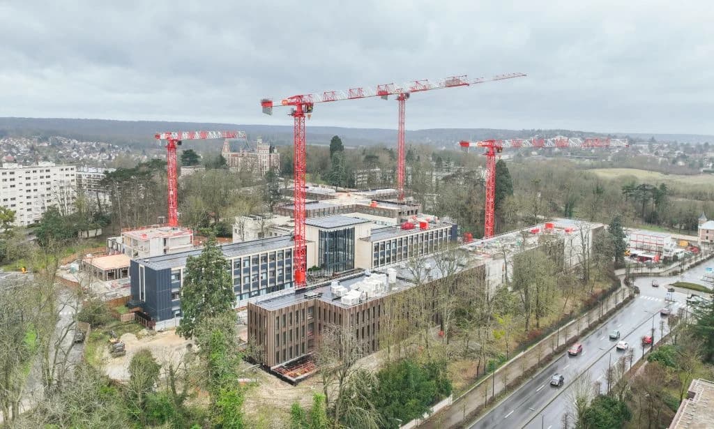 Vue aérienne d'un grand chantier de construction avec plusieurs grues rouges surplombant de nouveaux bâtiments et des structures existantes, entouré d'arbres et d'une route, avec un château en arrière-plan.
