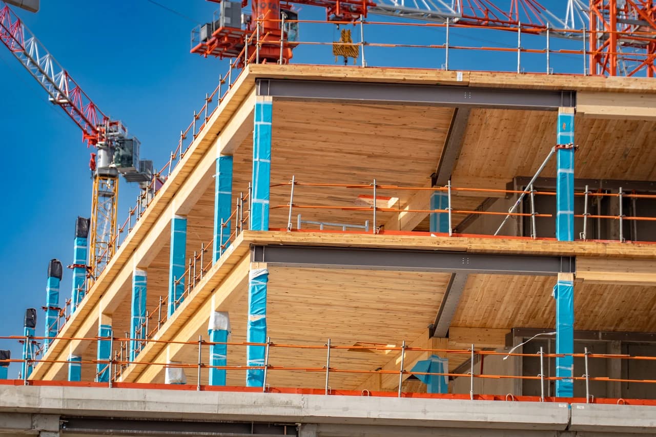 A multi-story building under construction, displaying light wood floors, dark steel structural beams, columns wrapped in bright blue plastic, and orange safety railings, all beneath a clear blue sky with distant cranes.