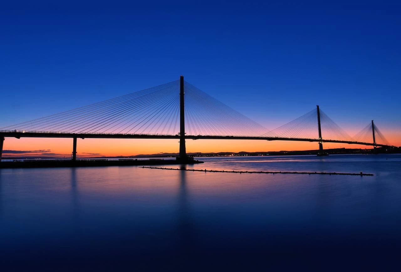 Un pont à haubans en silhouette au-dessus de l'eau calme, avec un ciel de coucher de soleil allant du bleu profond à l'orange vif, et des lumières lointaines sur la rive.