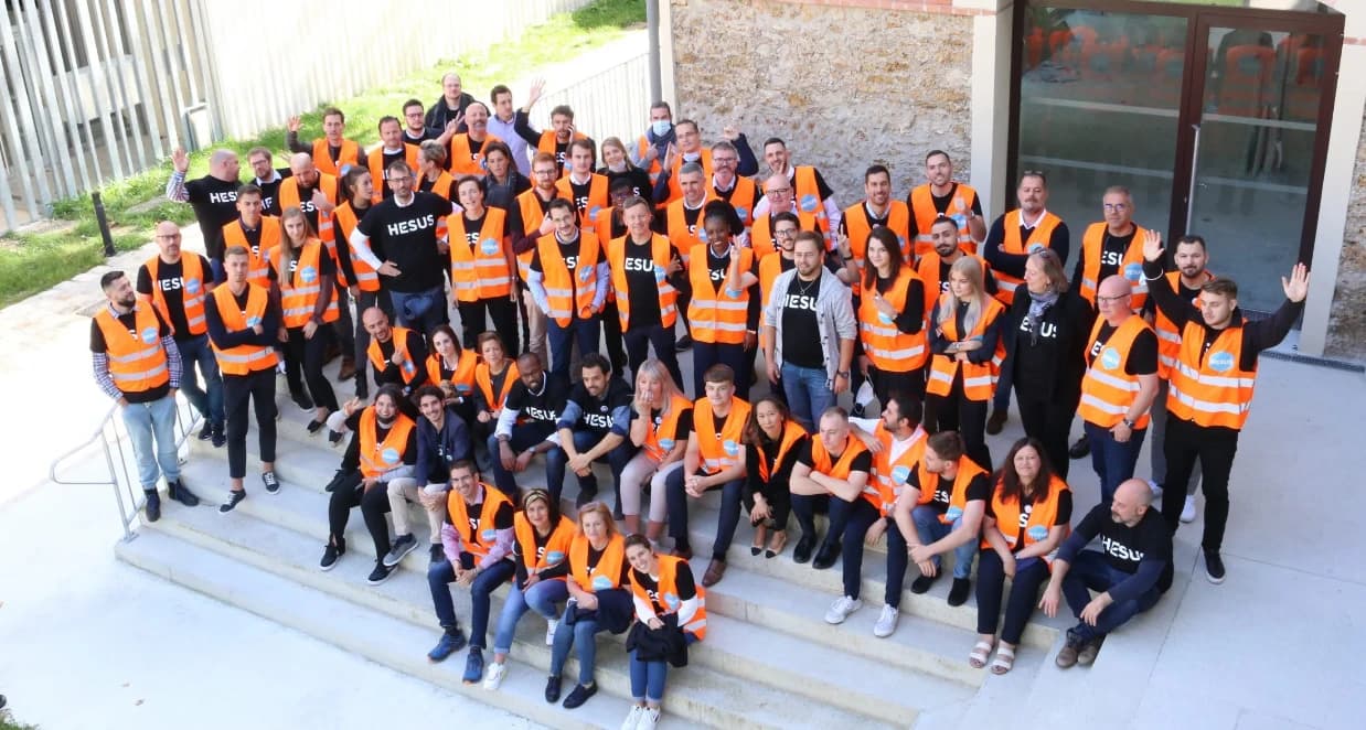 Un grand groupe de personnes diverses, vêtues de t-shirts noirs HESUS et de gilets de sécurité orange vif, posent ensemble et souriantes sur des escaliers extérieurs devant un bâtiment moderne.