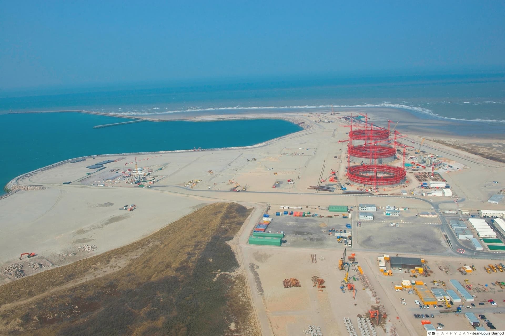 Vue aérienne d'un vaste chantier de construction industriel côtier, montrant trois grandes structures circulaires rouges en cours d'assemblage et un bassin portuaire artificiel turquoise relié à la mer.