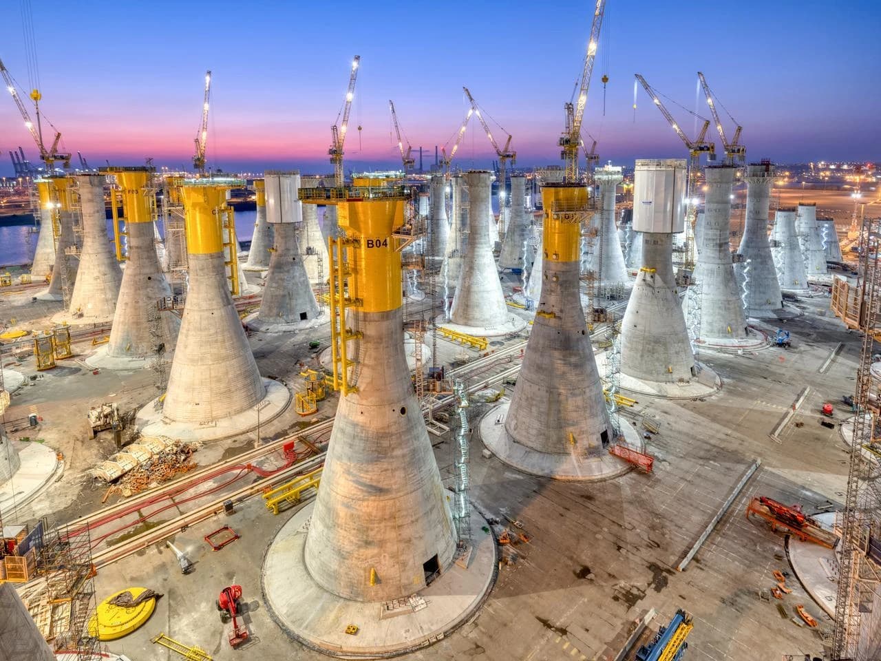 Une vaste installation portuaire au crépuscule présente des dizaines de bases de fondation coniques grises et jaunes pour éoliennes offshore. Des grues de construction sont visibles à travers le site sous un ciel aux teintes dégradées de bleu, rose et orange.