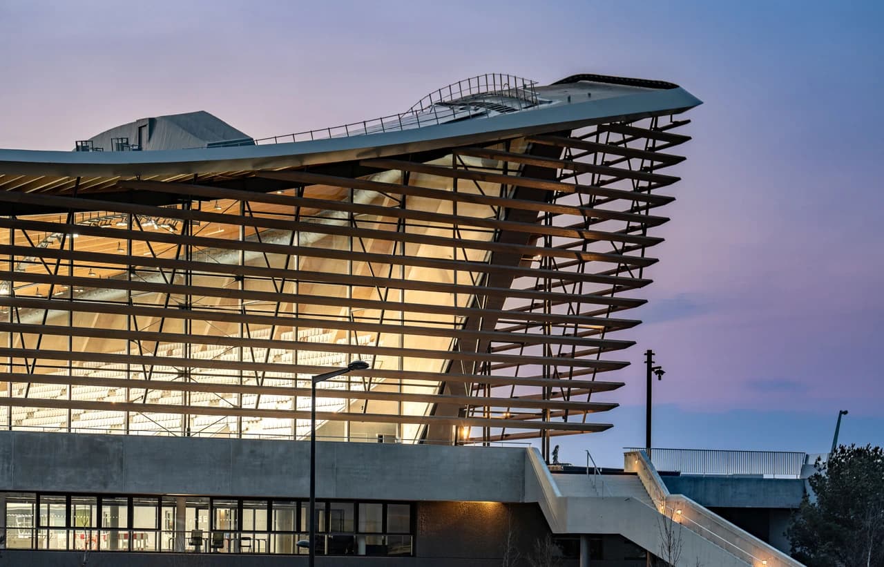 Bâtiment moderne avec un toit courbé et une façade en verre aux lattes horizontales, révélant un intérieur en bois chaleureusement éclairé. Le tout se dresse sous un ciel de crépuscule violet et bleu.