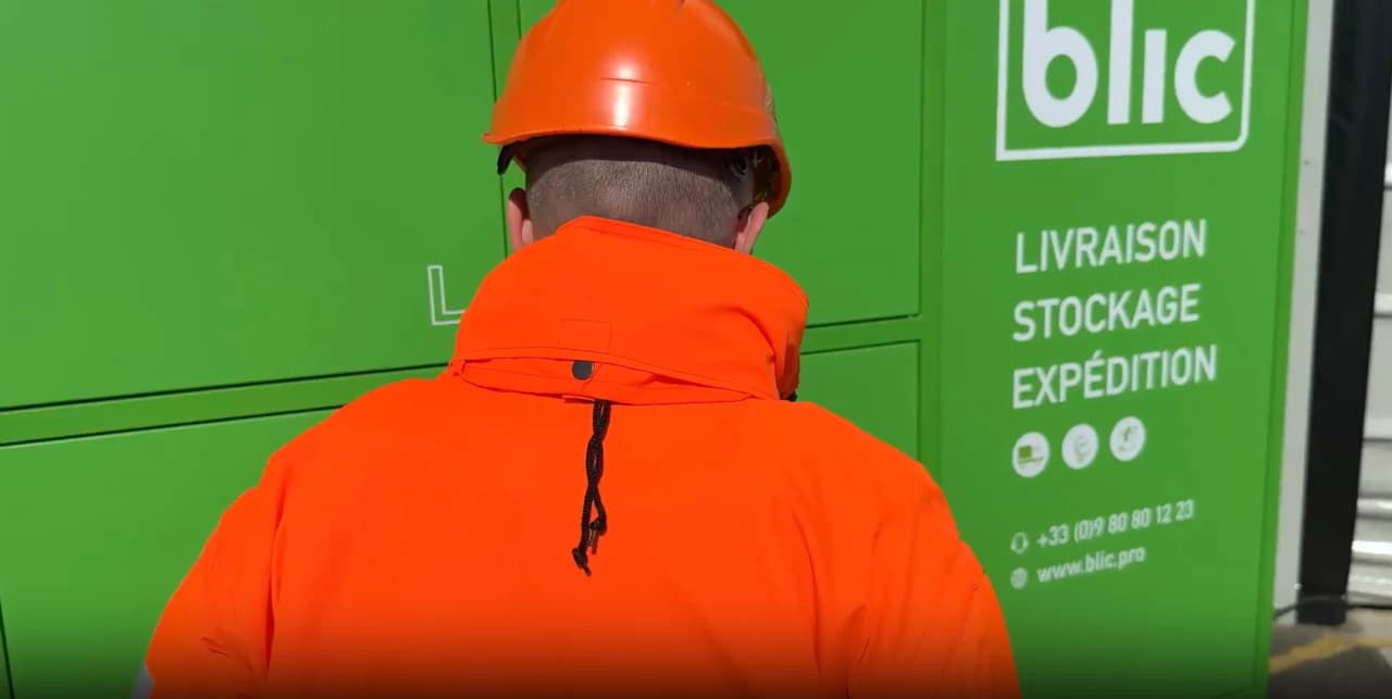 Une personne vêtue d'un casque et d'une veste de sécurité orange vif est vue de dos, face à un mur de casiers vert émeraude. Le mur affiche le logo "blic" et les services "LIVRAISON STOCKAGE EXPÉDITION".