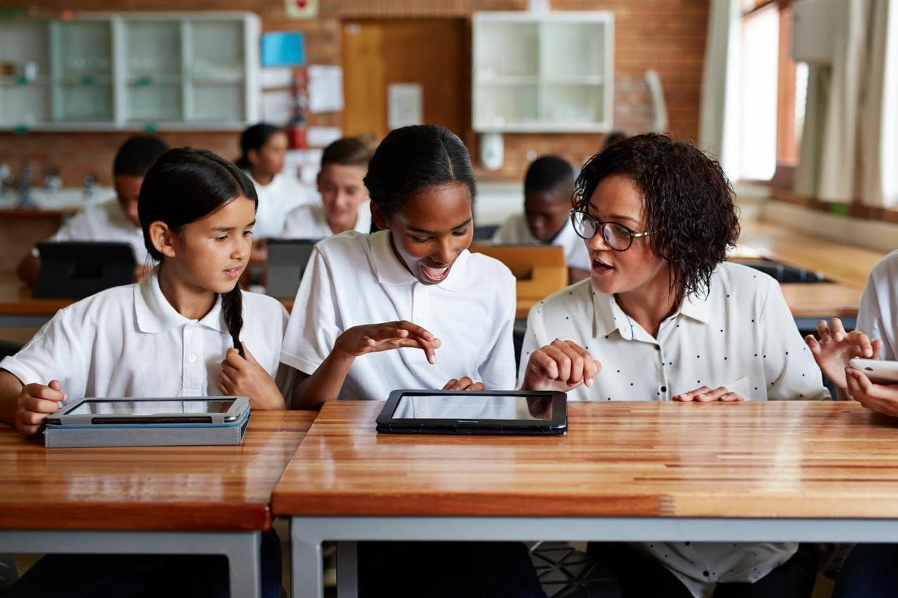 Deux écolières et une enseignante diverse travaillent avec des tablettes sur des pupitres en bois dans une salle de classe, l'enseignante guidant une élève souriante.