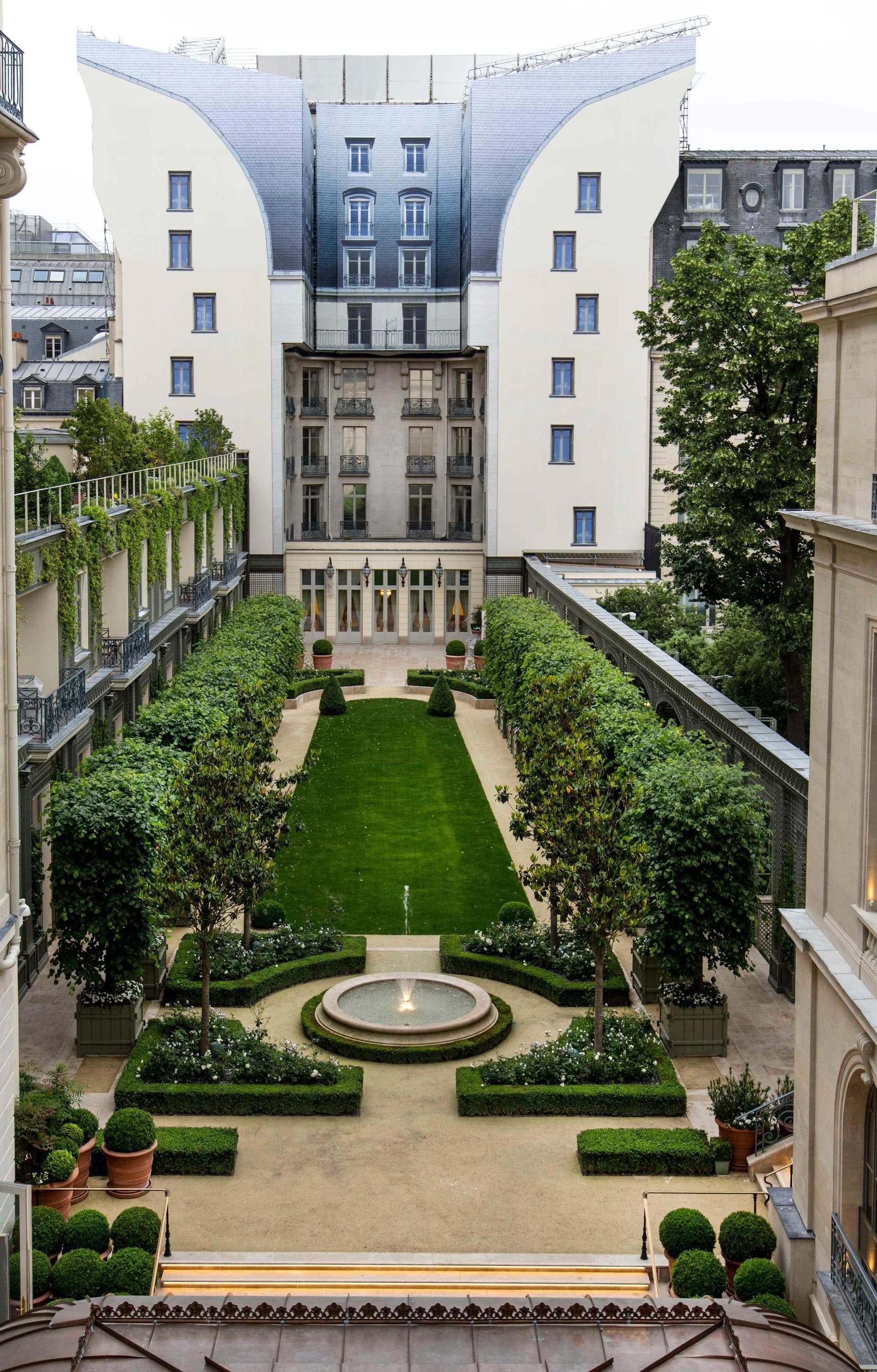 Un élégant jardin de cour symétrique présente une pelouse verte éclatante, une fontaine et des haies impeccablement taillées, encadré par des bâtiments classiques aux balcons ornés et une spectaculaire façade en ardoise grise courbée à l'arrière.