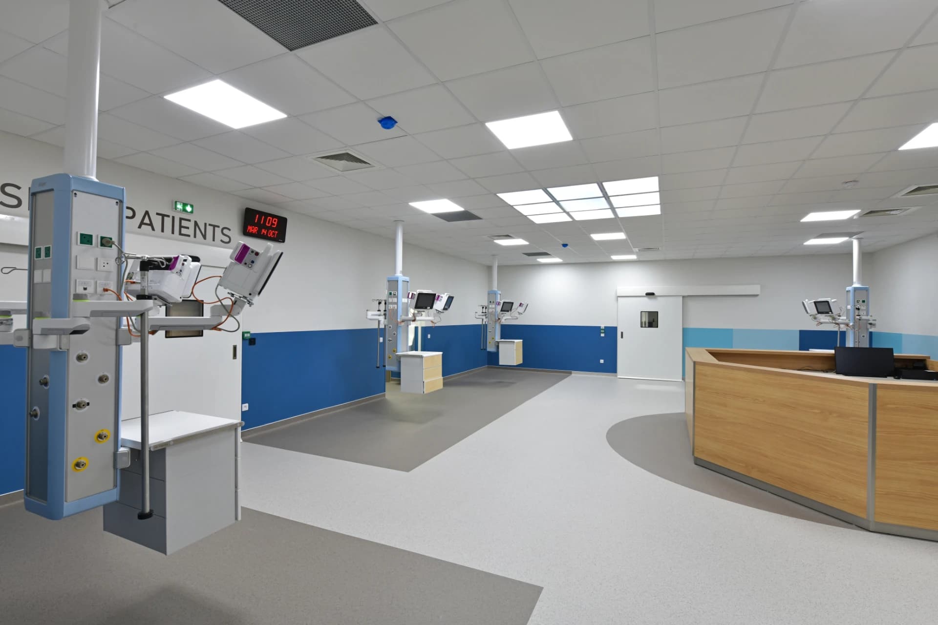 Salle d'hôpital moderne et lumineuse avec des murs blancs, une bande bleue, et un sol gris clair, équipée de plusieurs unités de soins suspendues au plafond et d'un comptoir d'accueil en bois. Un cadran numérique rouge sur le mur affiche l'heure et la date.