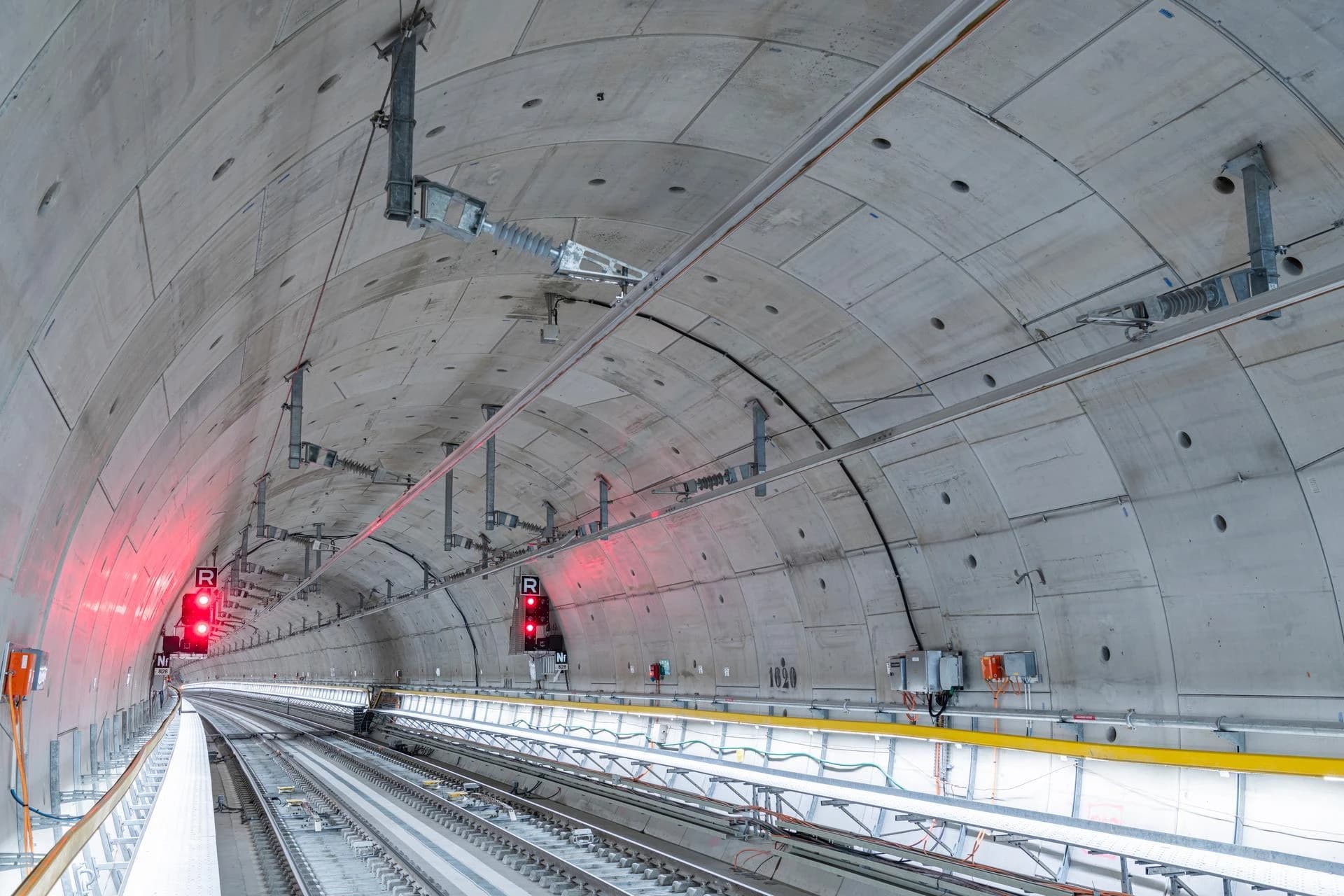 Un tunnel de train moderne en béton gris, courbé vers la droite dans la distance, présente deux voies ferrées et de nombreuses infrastructures électriques suspendues. Des feux de signalisation rouges vifs se détachent sur la paroi gauche, illuminant la scène.
