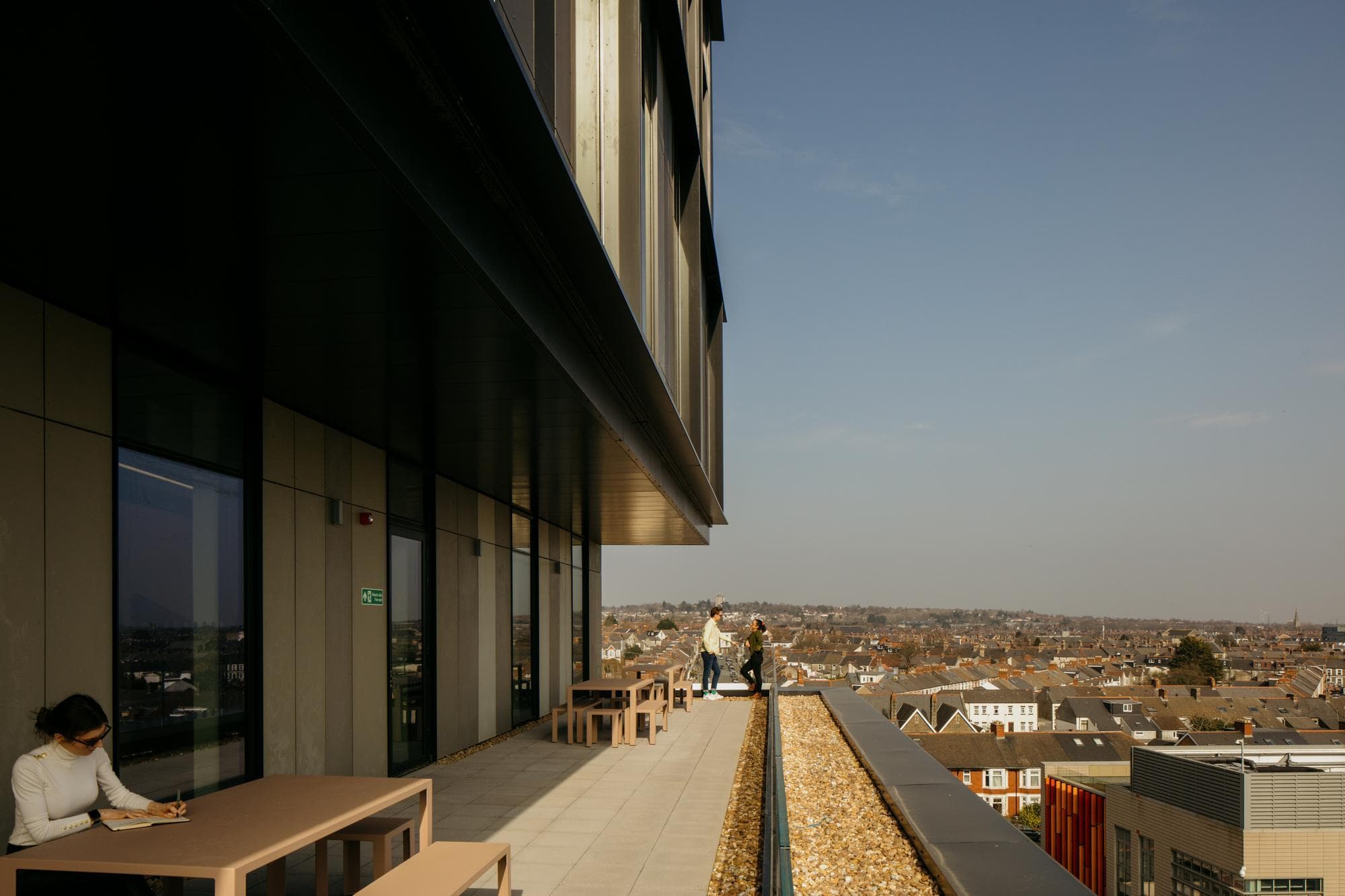 Une terrasse moderne avec des tables et bancs brun clair, où une femme en blanc écrit et deux personnes discutent, offre une vue étendue sur la ville sous un ciel bleu clair.