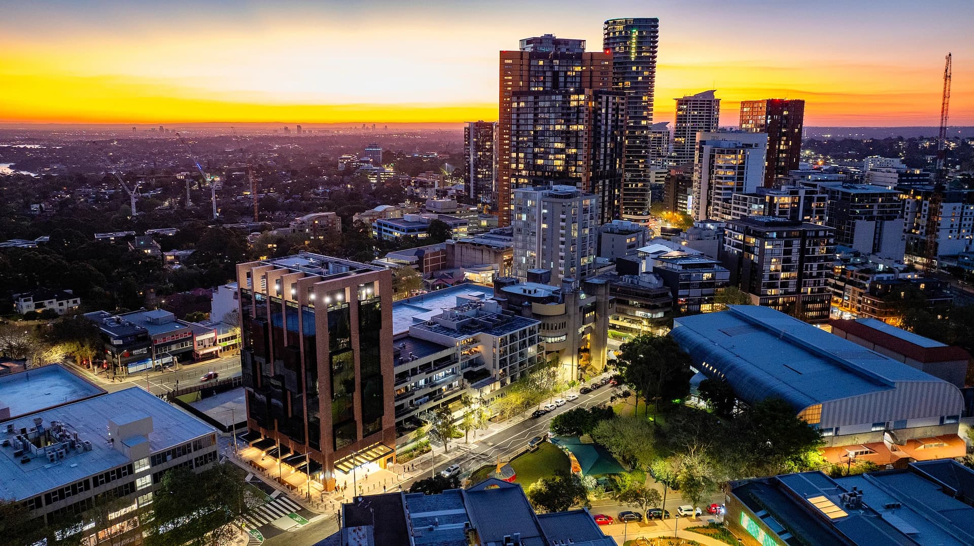 Vue aérienne d'un quartier urbain dense au coucher du soleil, montrant de nombreux immeubles de grande hauteur éclairés. Le ciel vibrant est teinté d'orange, de jaune et de violet au-dessus de l'horizon, contrastant avec les lumières chaudes de la ville.