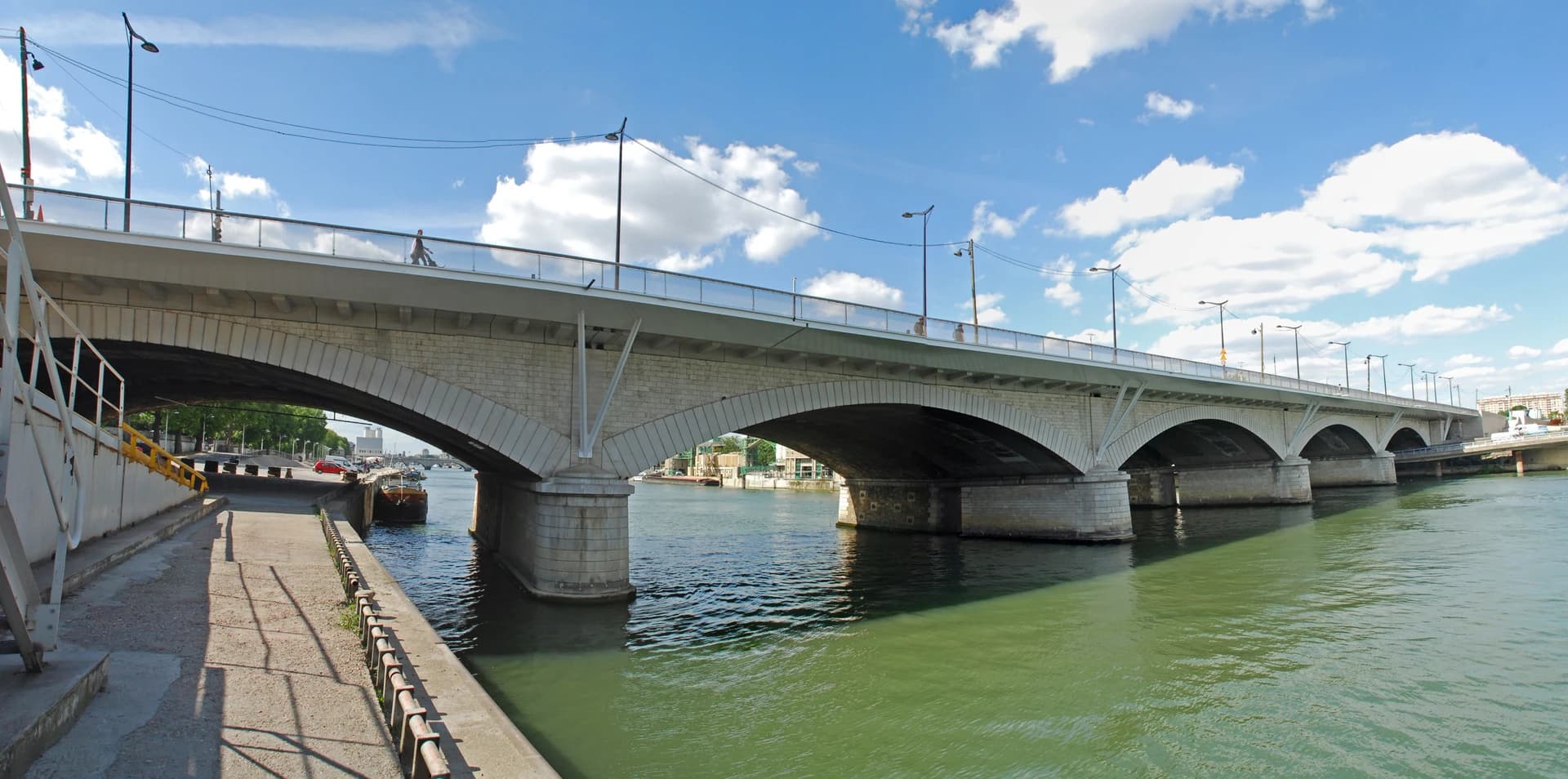 Un large pont en arc de pierre enjambe une rivière vert-bleu sous un ciel bleu clair nuageux. Des piétons traversent le pont moderne équipé de lampadaires, et une promenade bétonnée borde la rivière à gauche.