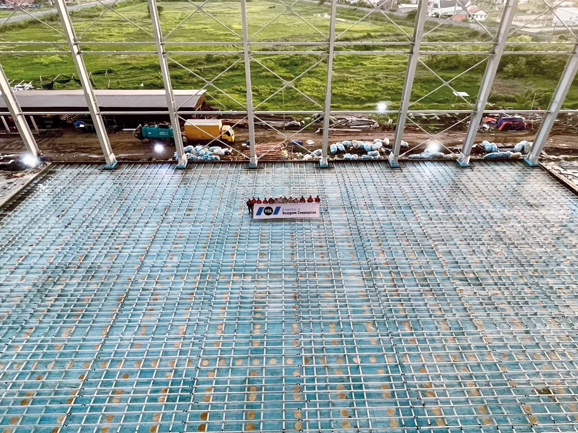 Un groupe d'environ dix personnes pose sur une vaste grille de ferraillage recouvrant une surface bleue, tenant une bannière "VSL A member of Bouygues Construction", avec en arrière-plan des véhicules de chantier et des champs verdoyants.