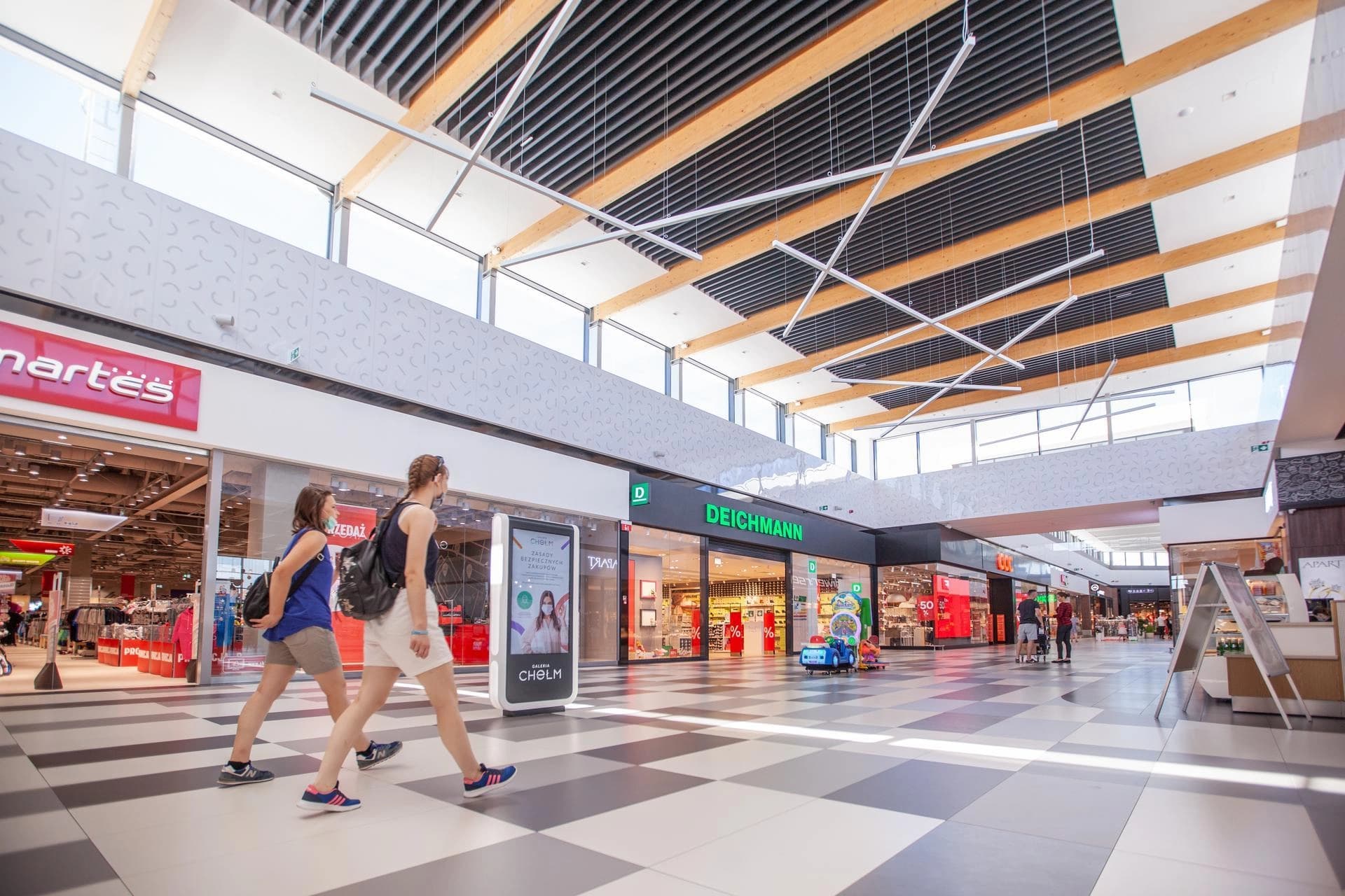 Intérieur d'un centre commercial moderne et lumineux avec un sol carrelé gris et blanc, où deux femmes marchent devant des boutiques telles que Martes et Deichmann, sous un haut plafond orné de poutres en bois et de luminaires linéaires.