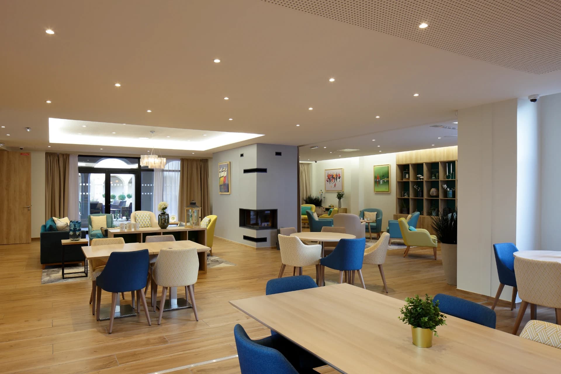 Vaste salle commune moderne d'une résidence, avec des tables de repas aux chaises bleues et beiges, un coin salon aux fauteuils colorés, une cheminée et de grandes fenêtres.