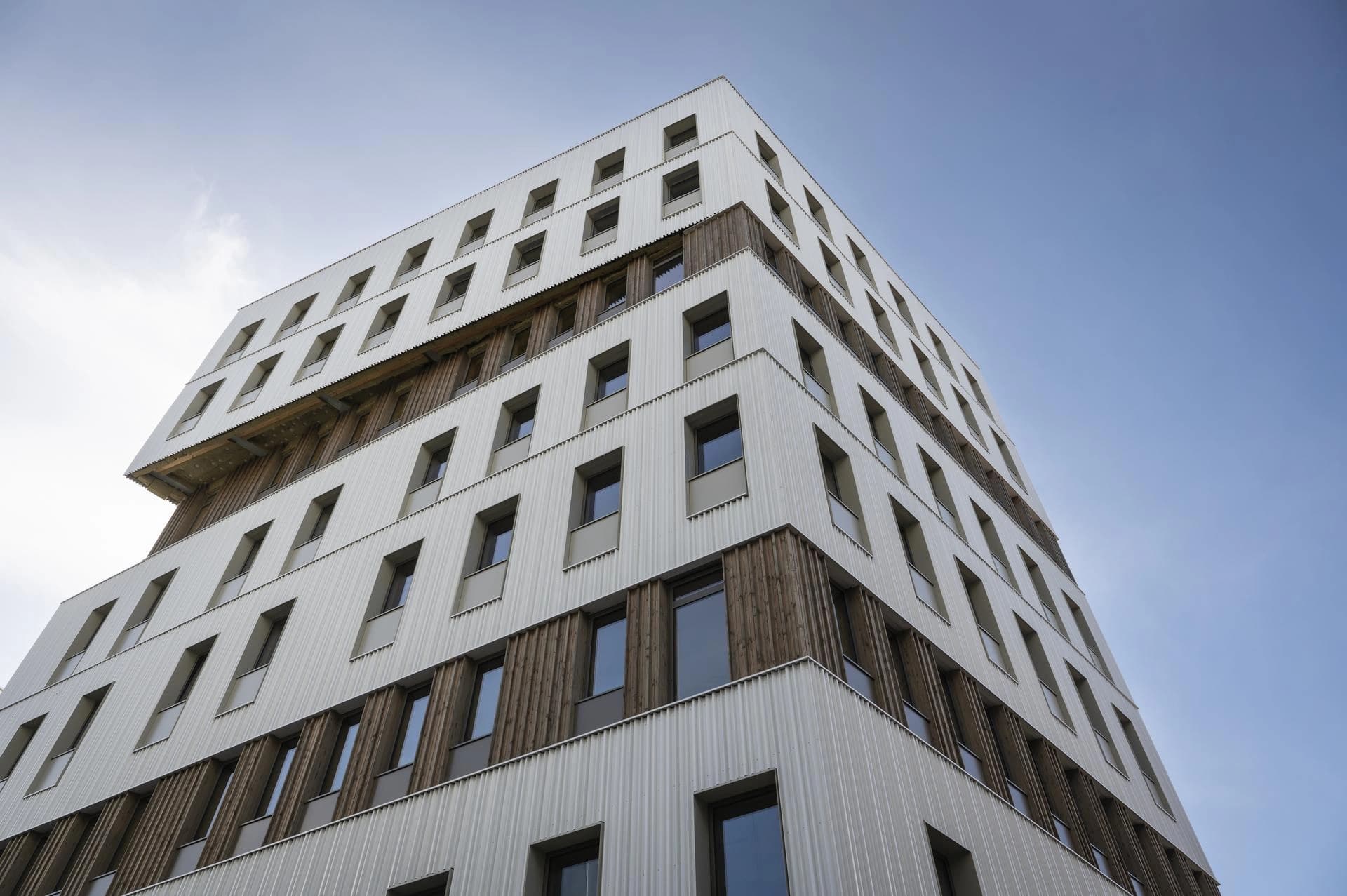 Bâtiment moderne à plusieurs étages avec revêtement blanc ondulé et panneaux de bois verticaux sous un ciel bleu clair.