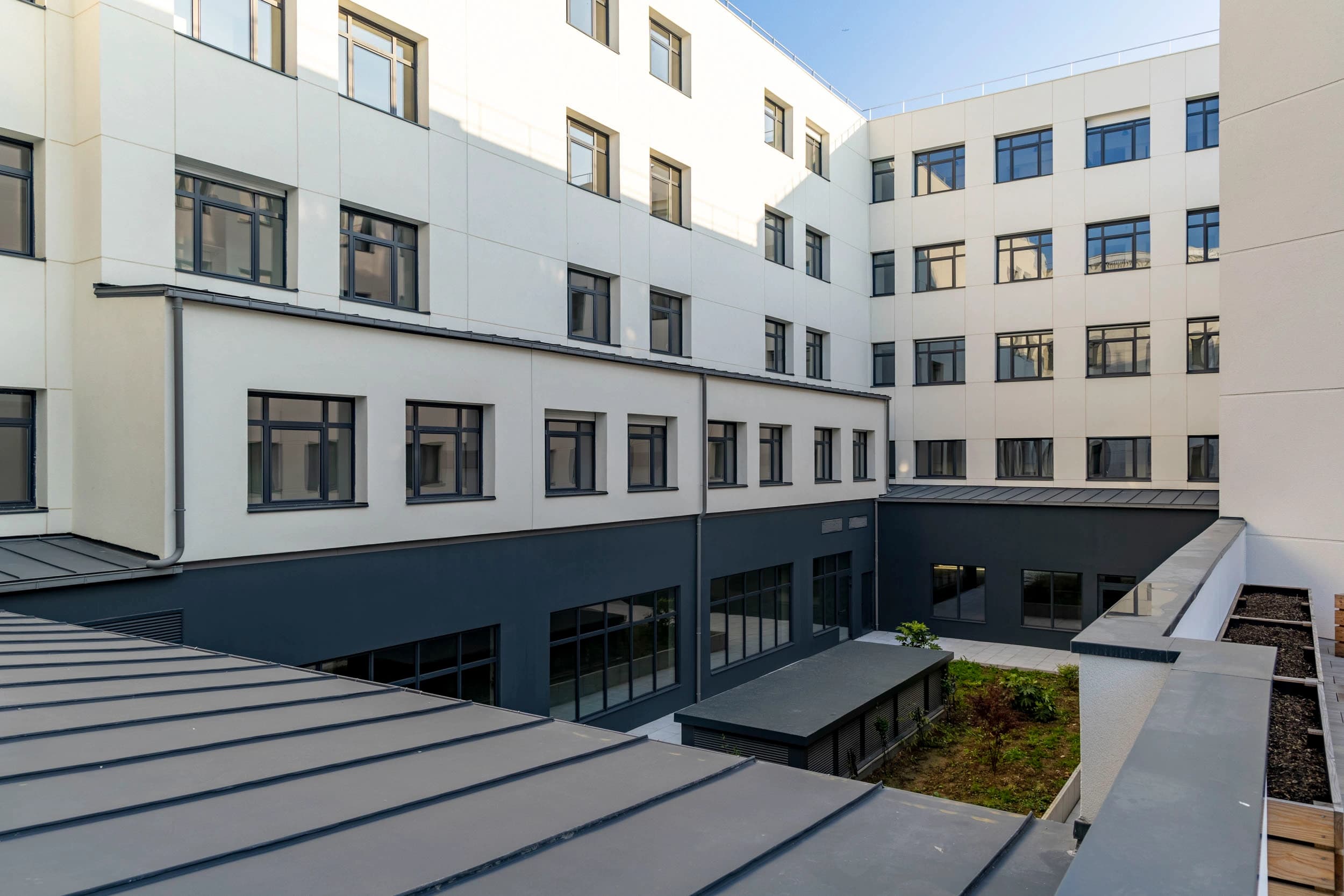 Cour intérieure d'un bâtiment moderne avec des façades blanc cassé et gris anthracite, de nombreuses fenêtres sombres, et un toit métallique gris en premier plan. Le patio comprend des zones pavées et de petits parterres de verdure sous un ciel bleu clair.