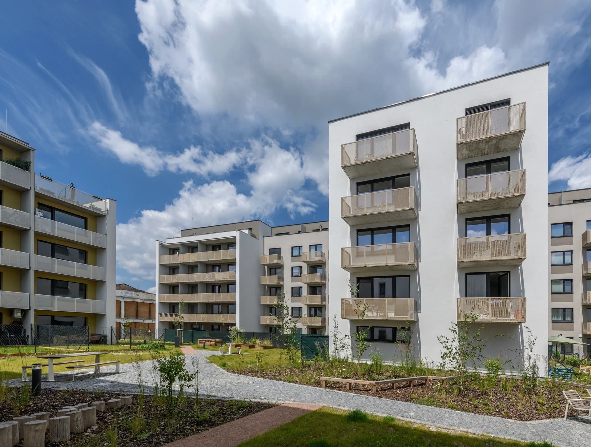 Plusieurs immeubles d'appartements modernes aux façades blanches et balcons perforés dorés encadrent un jardin paysager vert avec des chemins pavés et du mobilier de pique-nique, sous un ciel bleu parsemé de nuages blancs.