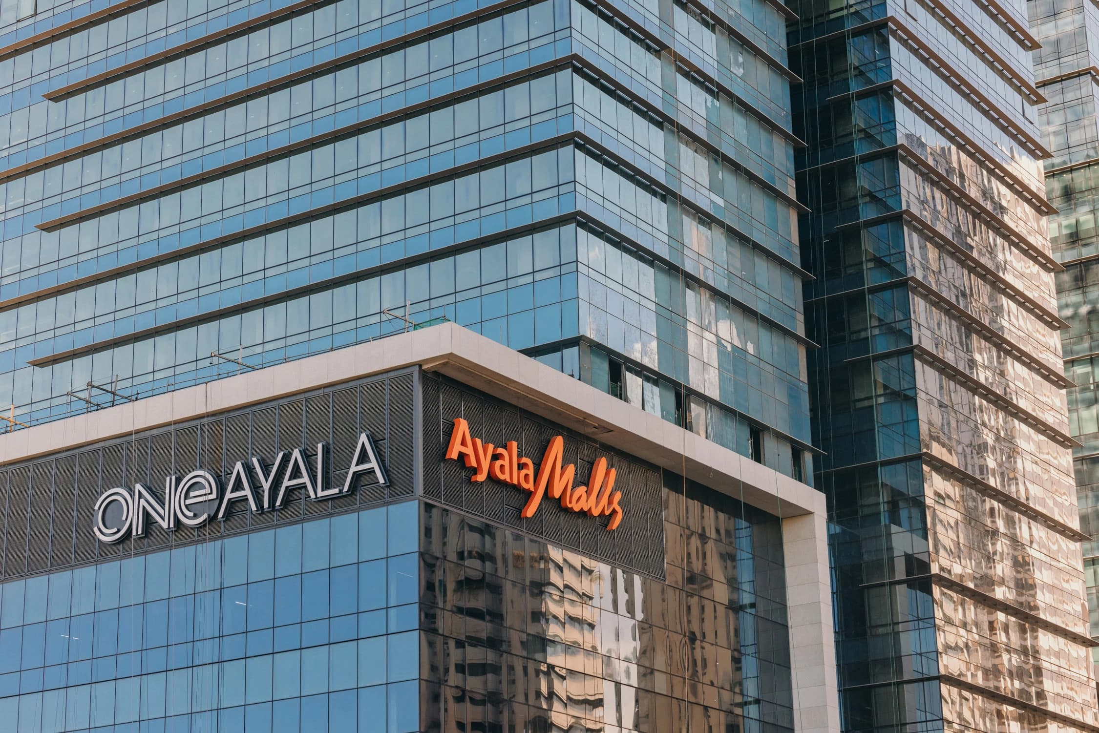 Façade d'un immeuble moderne entièrement vitré, avec les enseignes "ONE AYALA" en blanc et "Ayala Malls" en orange vif sur un bandeau gris foncé.