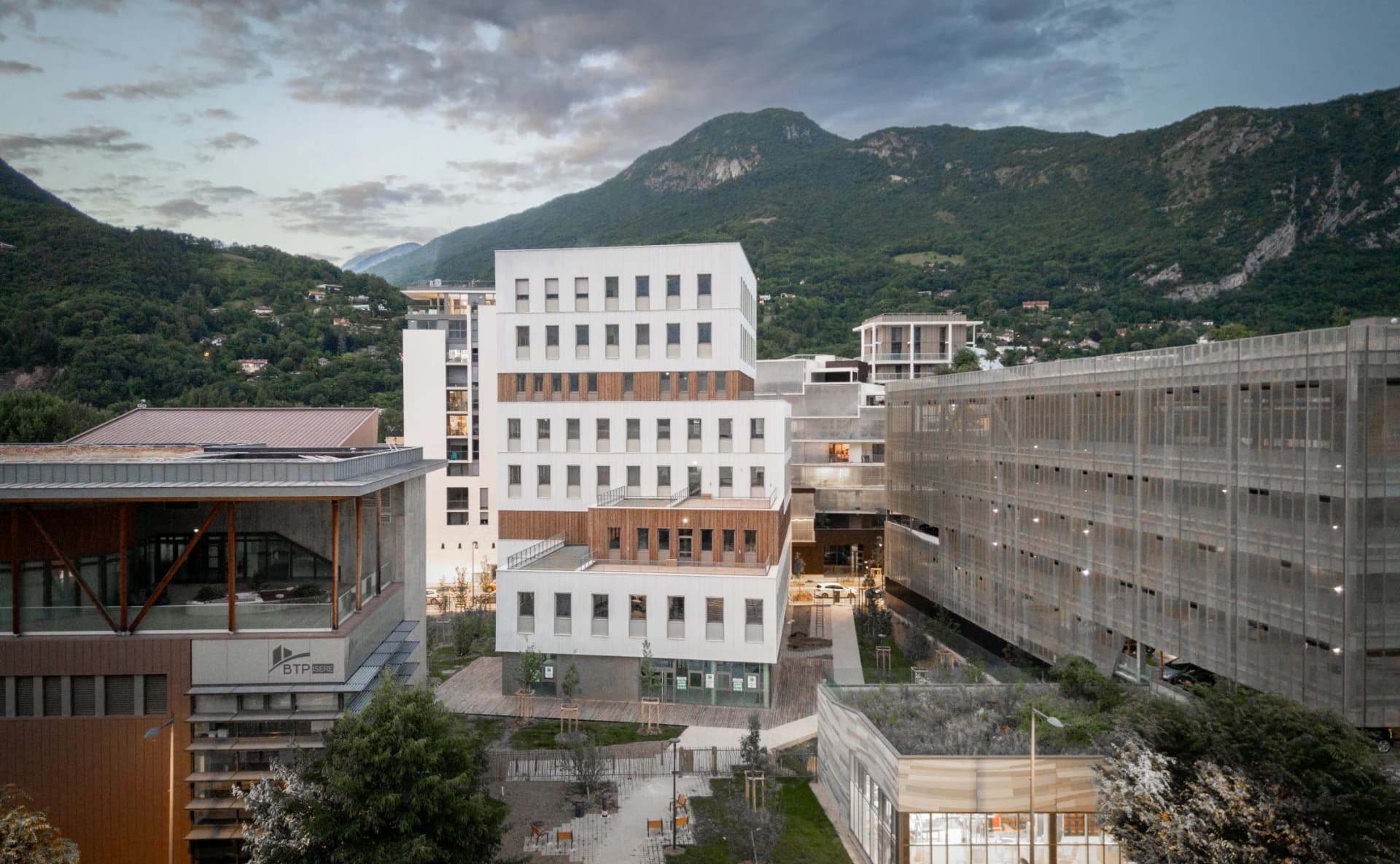 Un complexe architectural moderne, composé d'un grand bâtiment blanc avec des accents de bois au centre, d'un parking à façade grillagée, et d'autres constructions contemporaines, est intégré dans un paysage montagneux verdoyant sous un ciel nuageux.