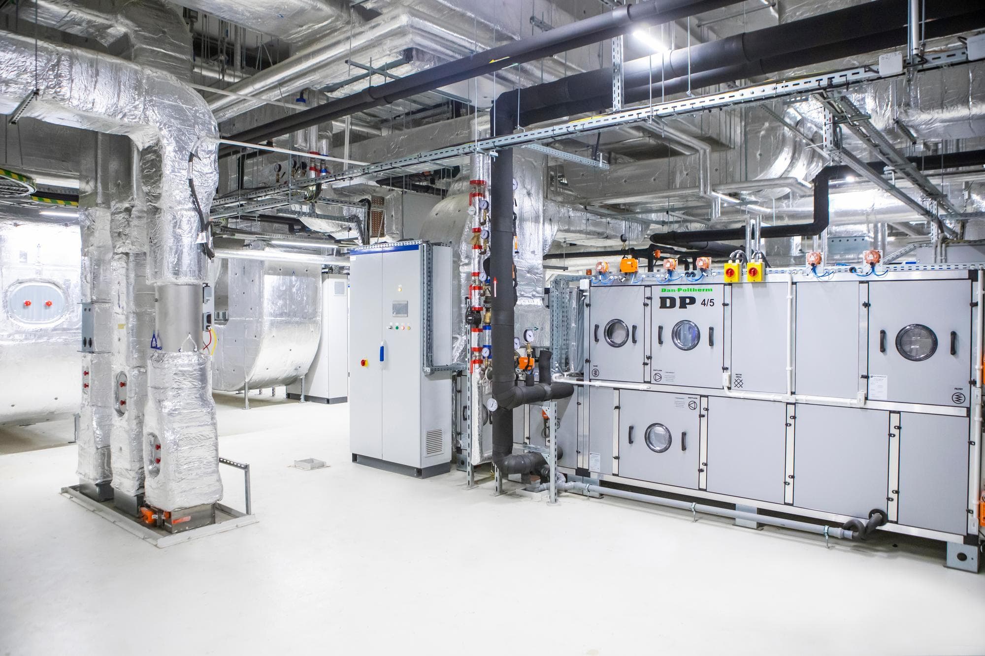 Une salle technique moderne abrite une grande unité de traitement d'air grise Dan-Polltherm, entourée d'un réseau complexe de conduits isolés argentés et de tuyaux noirs, sur un sol blanc impeccable.
