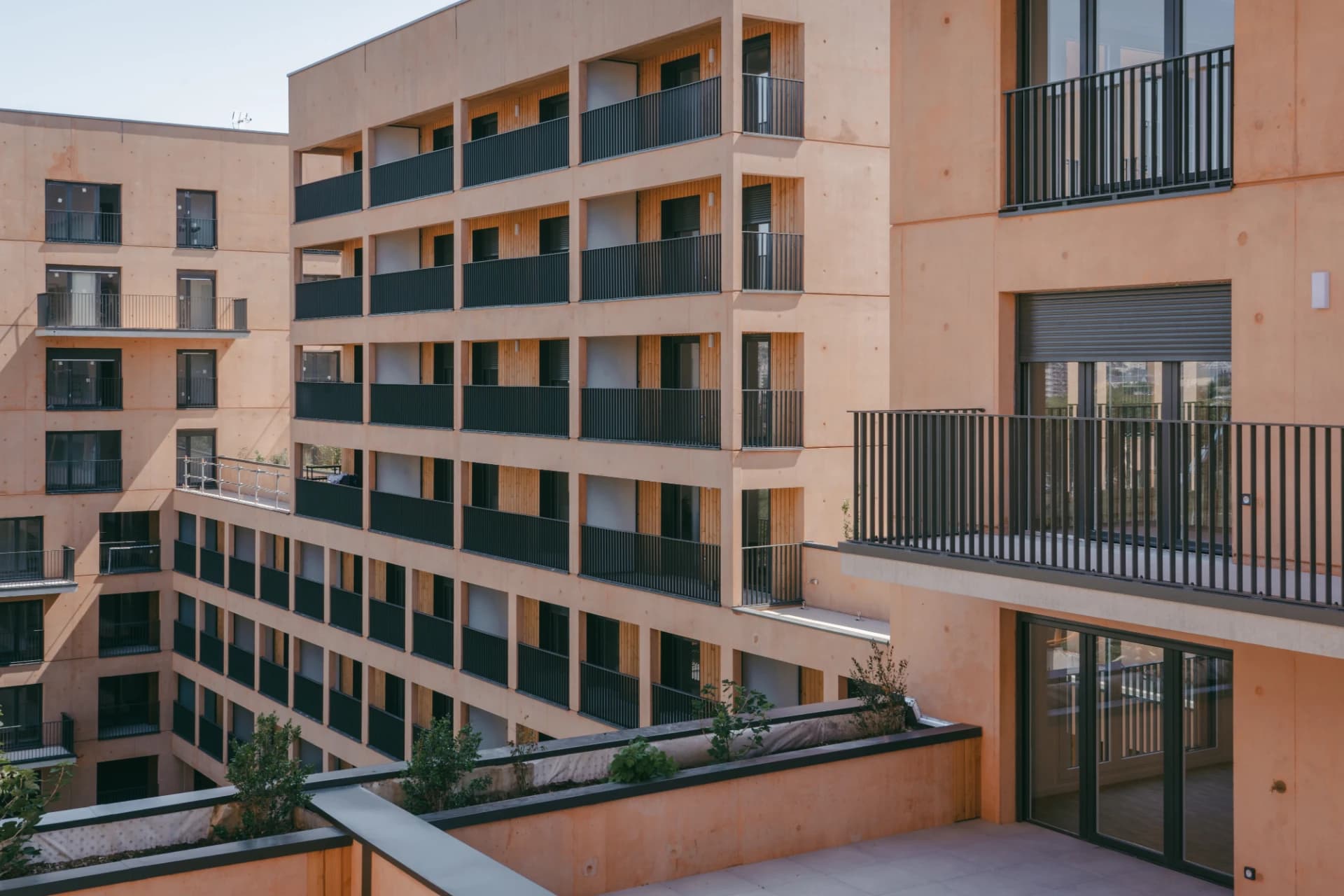 Plusieurs bâtiments résidentiels modernes aux façades couleur terre cuite ou pêche, avec de nombreux balcons à garde-corps sombres. Des fenêtres et portes-fenêtres foncées sont visibles, complétées par des jardinières avec de la végétation sur les terrasses.