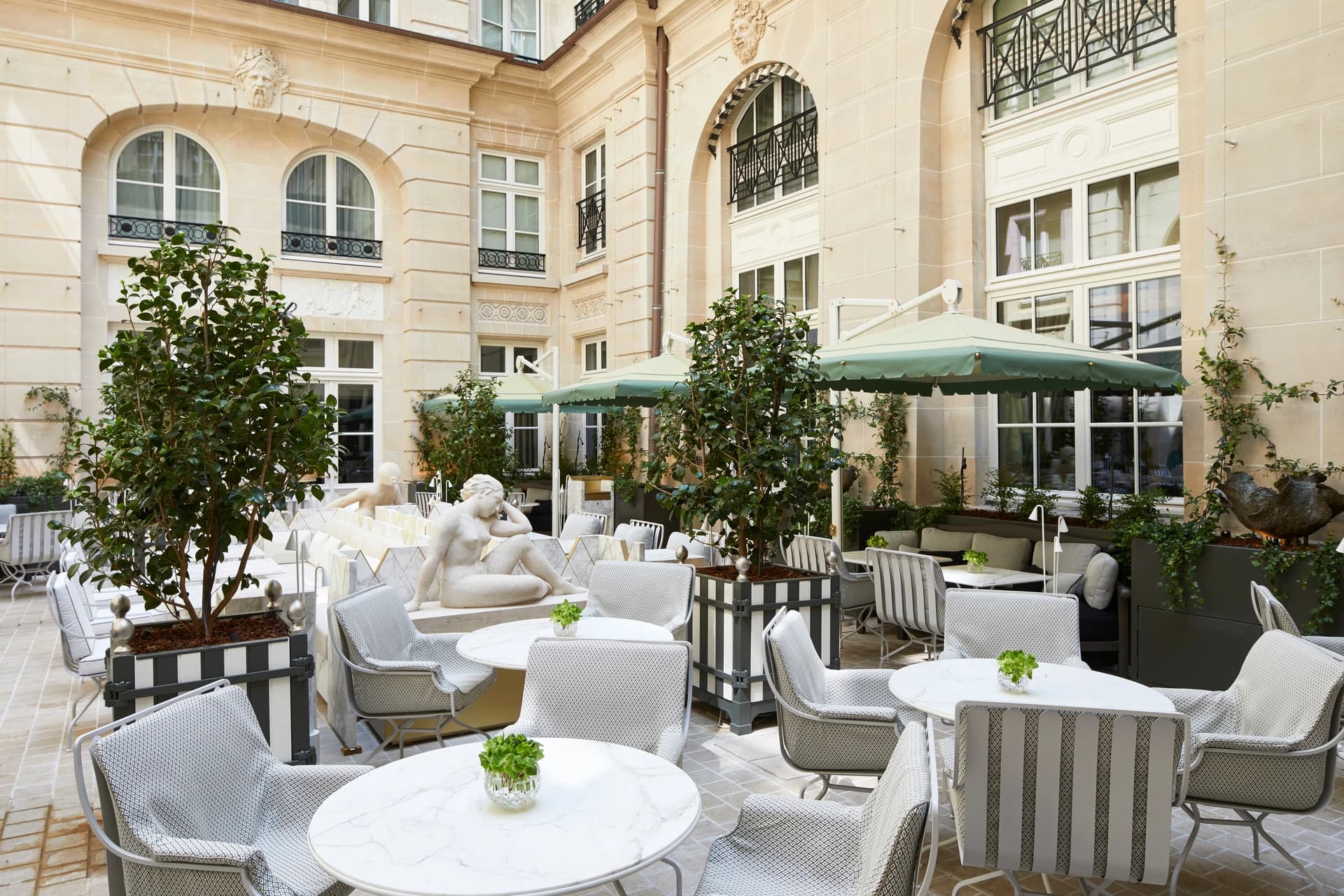 Un patio extérieur luxueux avec des tables en marbre blanc, des chaises grises et blanches, et des parasols verts sous une élégante façade en pierre claire ornée de plantes et de statues classiques.