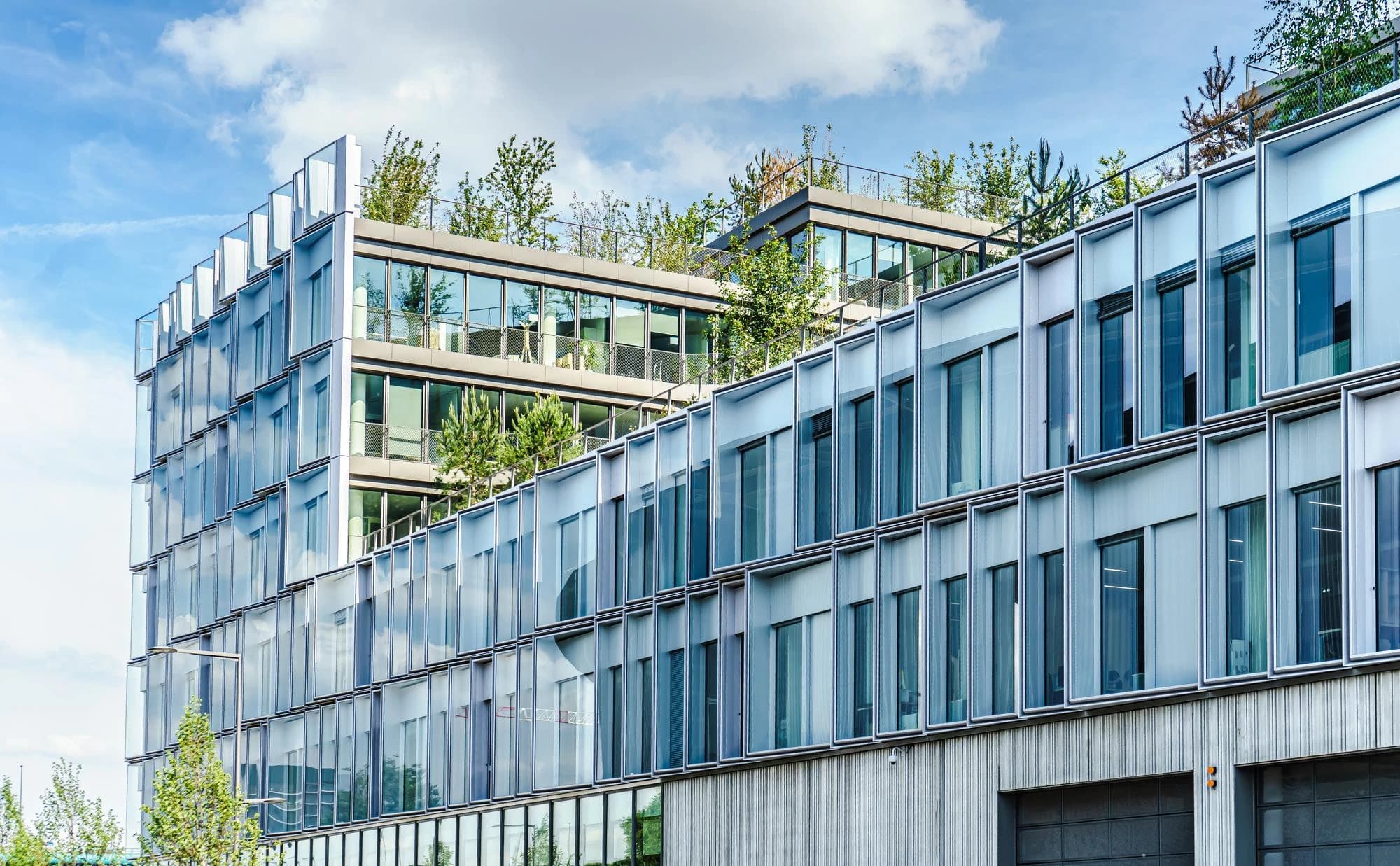 Bâtiment de bureaux moderne avec une façade de verre et des toits végétalisés luxuriants, reflétant le ciel bleu parsemé de nuages blancs.