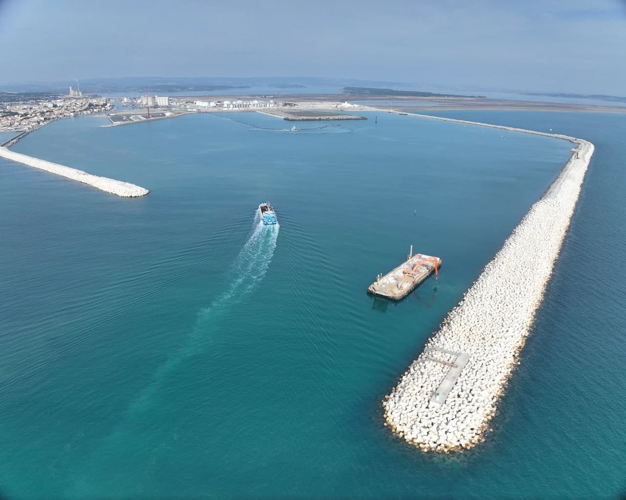 Vue aérienne d'un grand port aux eaux turquoise, où un ferry bleu et blanc entre entre de longues digues en béton blanc, près desquelles une barge de construction est amarrée.