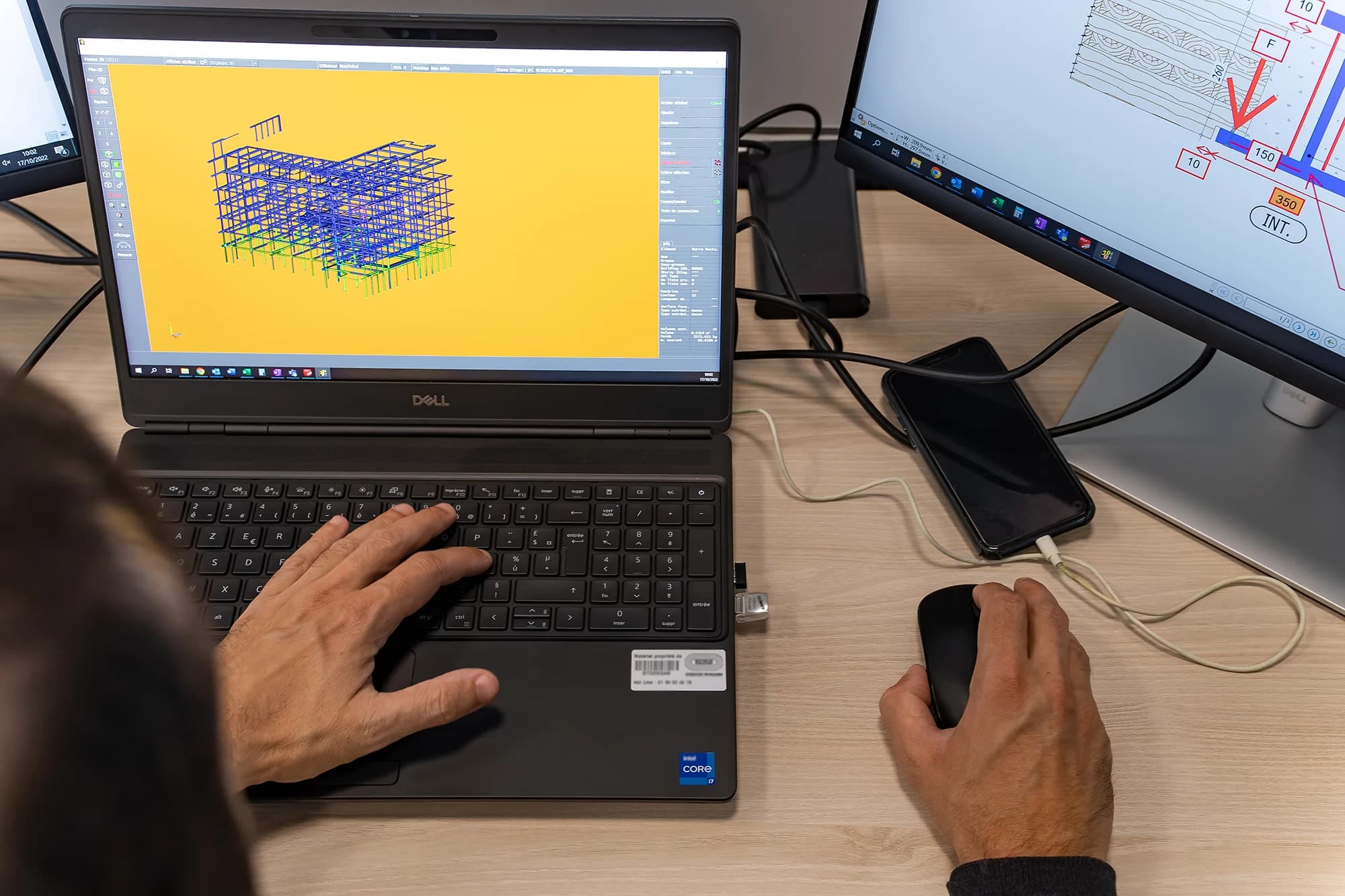 Vue plongeante sur une personne travaillant à un bureau, avec un ordinateur portable affichant un modèle 3D de structure de bâtiment sur fond jaune et un second moniteur montrant des plans techniques, les mains sur le clavier et la souris.