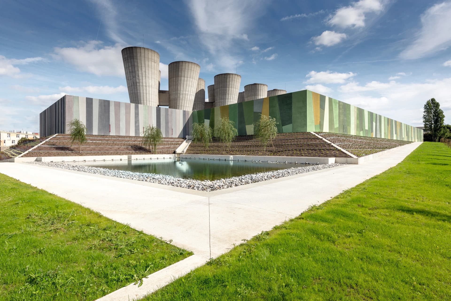 Un bâtiment moderne avec une façade de panneaux verticaux gris et pastels transitionnant vers des motifs géométriques verts, surmonté de plusieurs tours de refroidissement grises. Au premier plan, un étang triangulaire est entouré d'herbe verte et d'un chemin en béton menant à un talus paysager sous un ciel bleu nuageux.