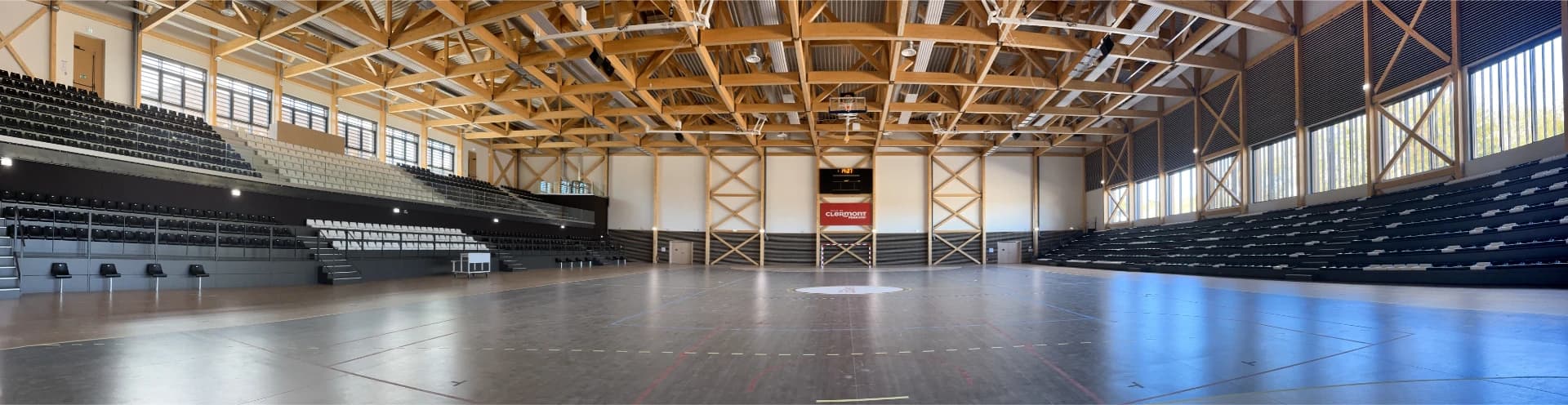 Vue panoramique d'un gymnase moderne et lumineux, caractérisé par une imposante charpente apparente en bois clair et des gradins sombres sur les côtés. Un terrain de sport polyvalent occupe le centre, et un tableau d'affichage montre l'heure et le logo "Ville de Clermont Ferrand".