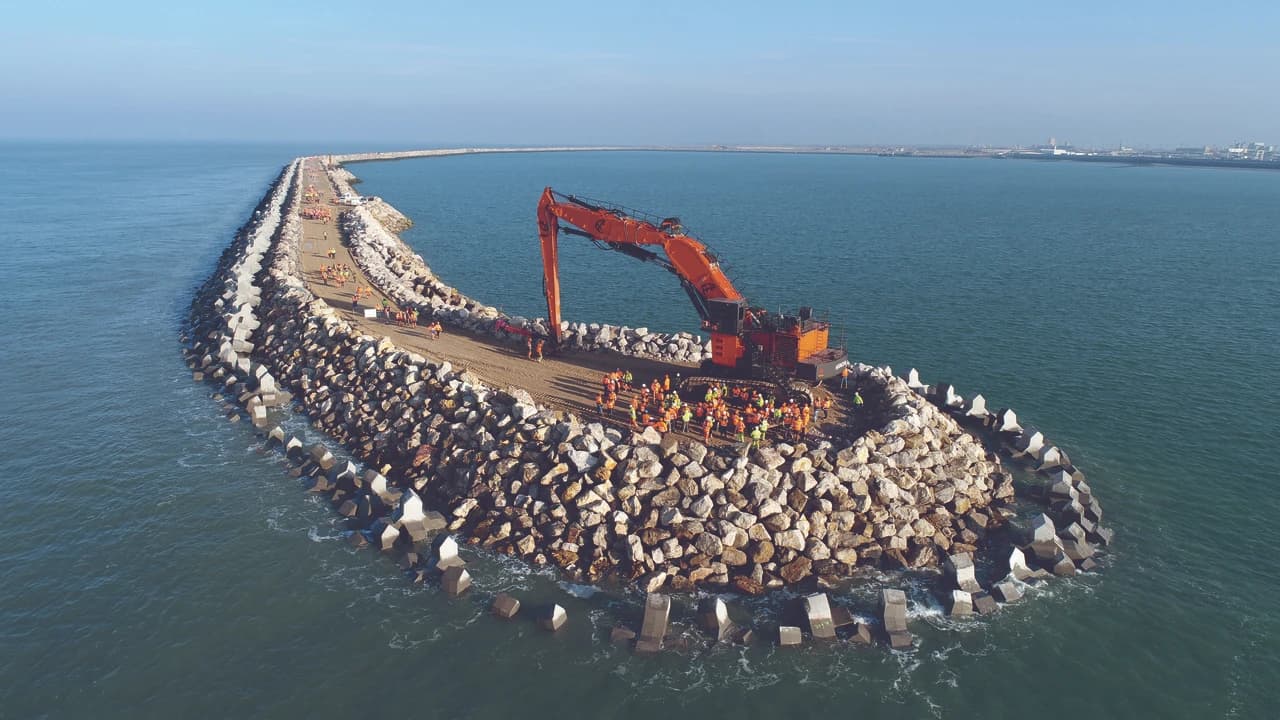 Vue aérienne d'un brise-lames en construction, une longue digue courbée de roches et de blocs de béton dans la mer bleue, où une grande pelle mécanique orange est entourée d'ouvriers en gilets de sécurité, avec un port visible au loin.