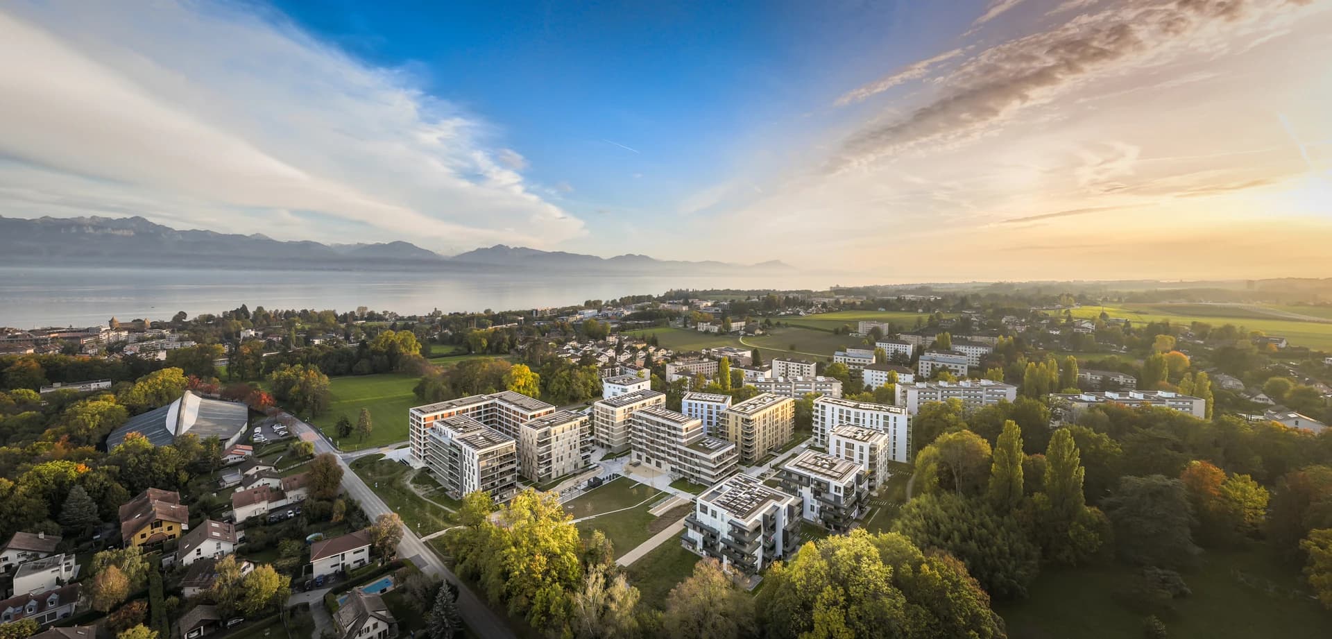 Vue aérienne d'un complexe résidentiel moderne aux façades claires et toits solaires, entouré de verdure et d'arbres aux couleurs automnales, avec un grand lac et des montagnes en arrière-plan sous un coucher de soleil éclatant.