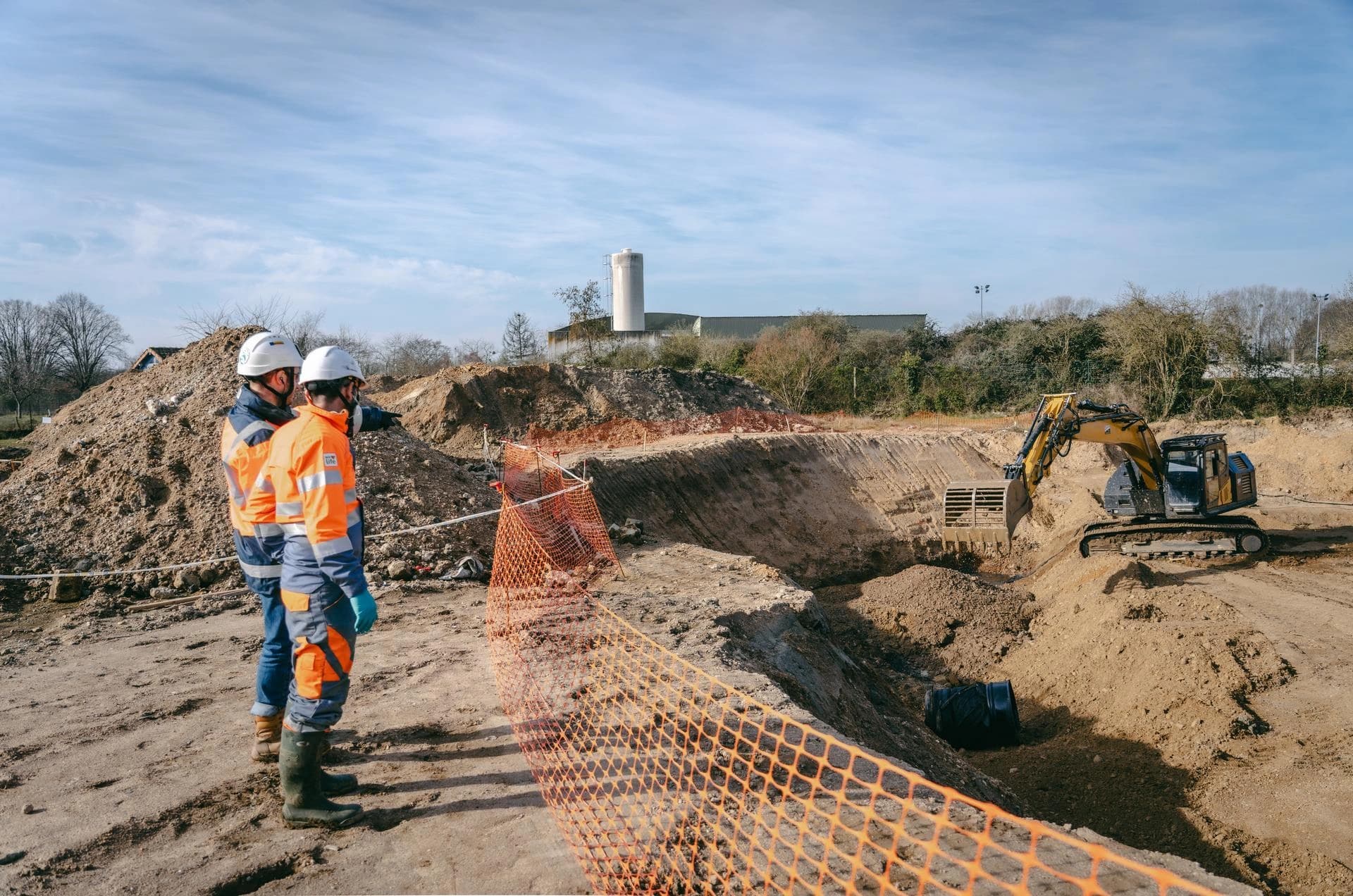 Deux ouvriers vêtus de gilets orange et casques blancs supervisent un chantier de terrassement où une pelleteuse creuse une grande fosse. Le site présente des monticules de terre, des clôtures de sécurité orange et, à l'arrière-plan, des arbres et des bâtiments sous un ciel bleu clair.