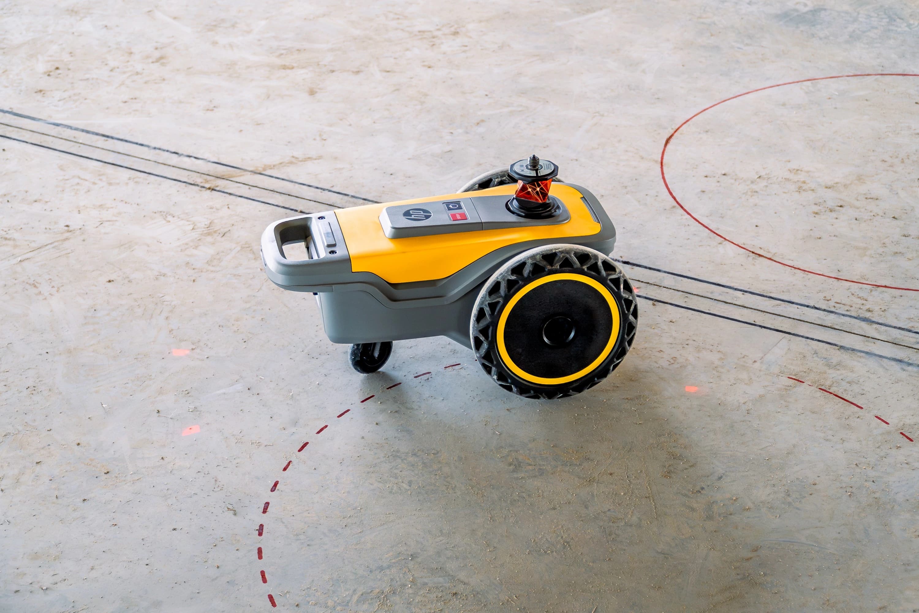 Un robot de marquage jaune et gris, avec un logo HP et un prisme de mesure rouge sur le dessus, est posé sur un sol clair parsemé de lignes noires et rouges.