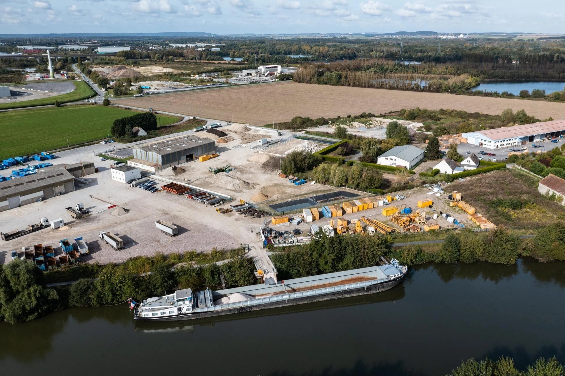 Vue aérienne d'un site industriel fluvial actif, montrant un long chaland chargé de granulats amarré à un quai. Le site est pourvu d'entrepôts, de camions, de piles de matériaux et de conteneurs jaunes, bordé par des zones vertes et des champs agricoles sous un ciel partiellement nuageux.