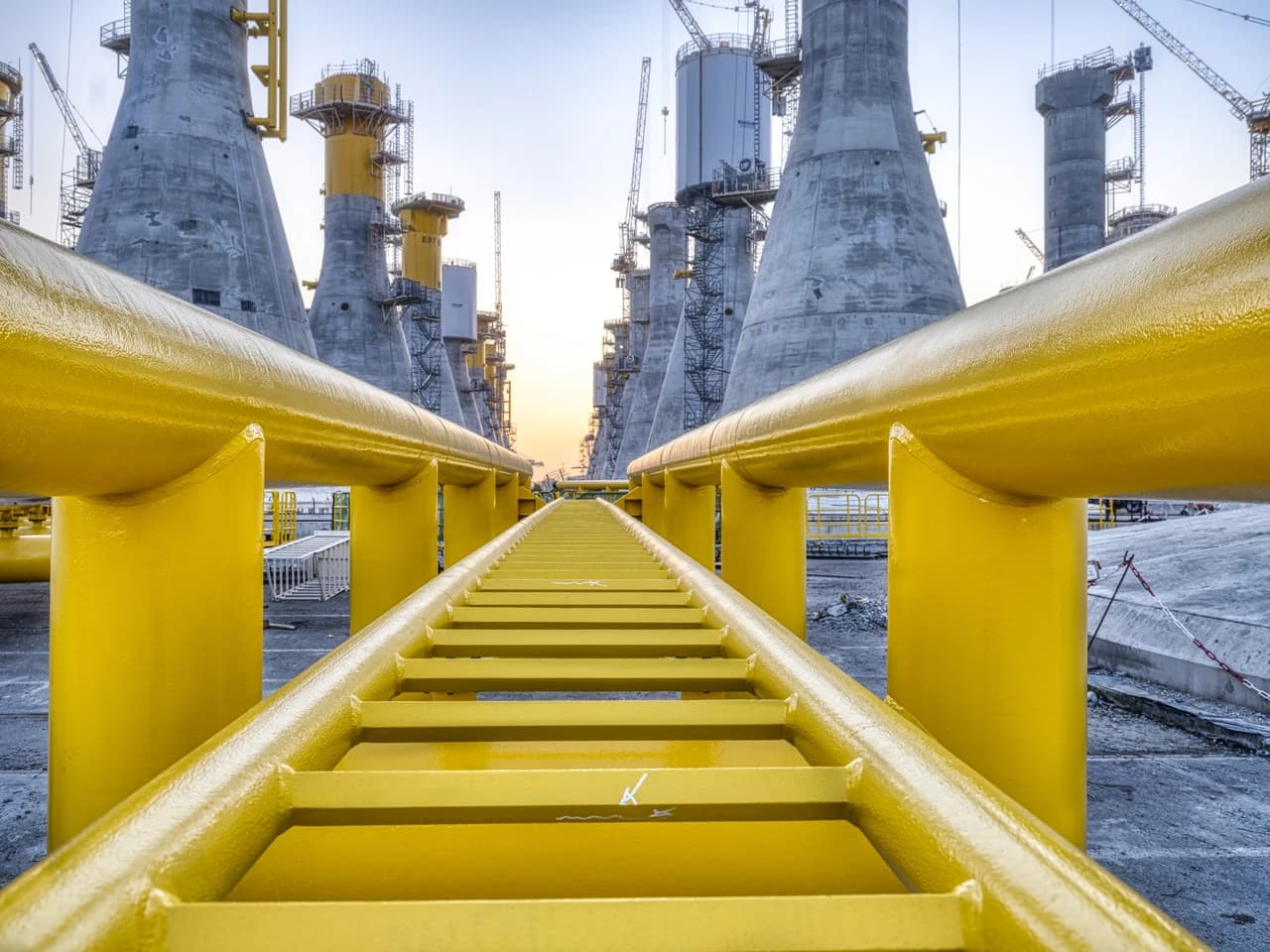 Une allée ou tuyauterie industrielle jaune vif et brillante s'étend au premier plan, encadrant une vaste zone de construction avec d'imposantes structures coniques en béton gris et des grues sous un ciel clair.
