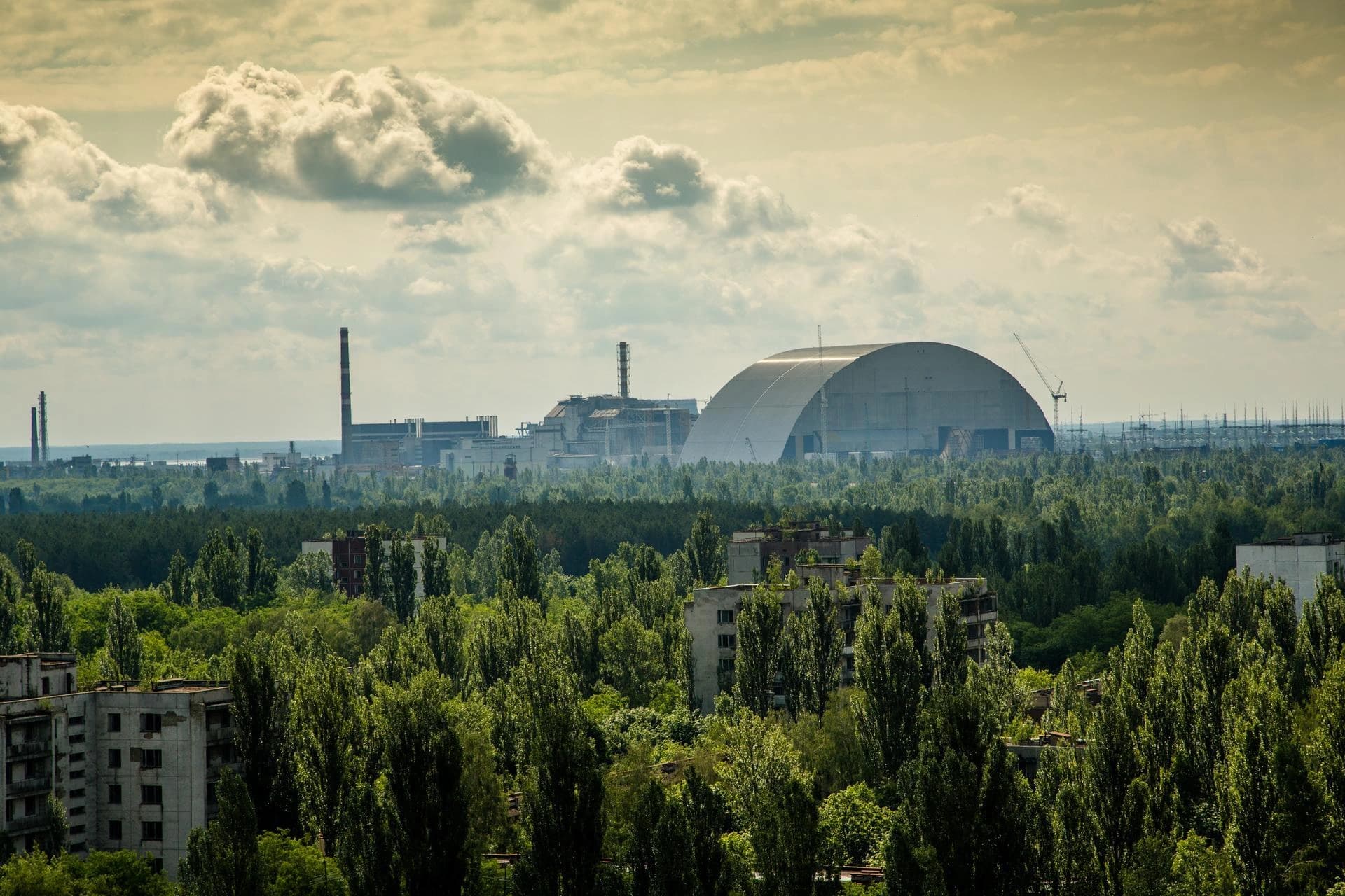 Une vue lointaine du nouveau sarcophage arqué gris de Tchernobyl et des bâtiments de la centrale, sous un ciel nuageux. Au premier plan, de denses arbres verts envahissent des bâtiments résidentiels abandonnés.