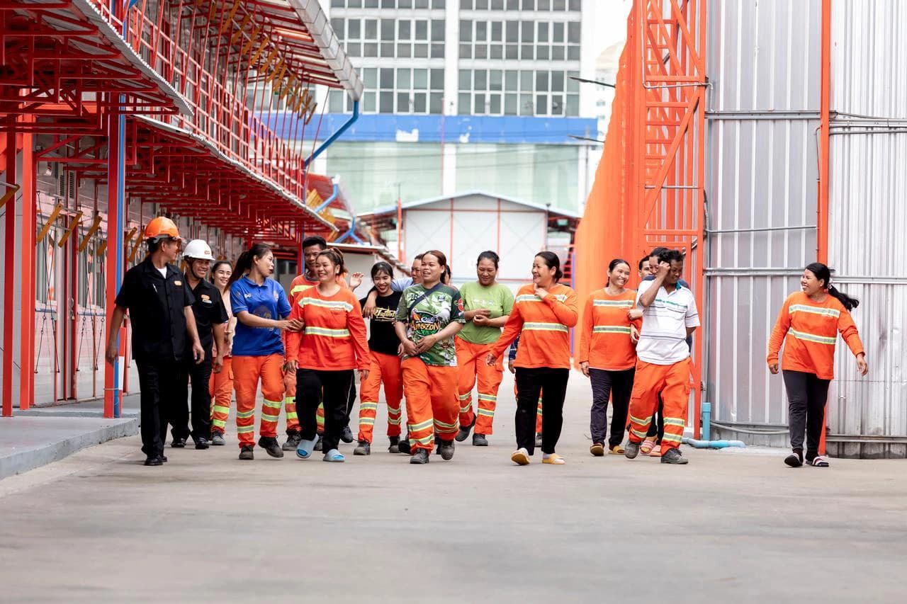 Un groupe diversifié de travailleurs, principalement des femmes portant des uniformes de travail orange, marche ensemble et sourit dans une usine avec des structures métalliques rouges.