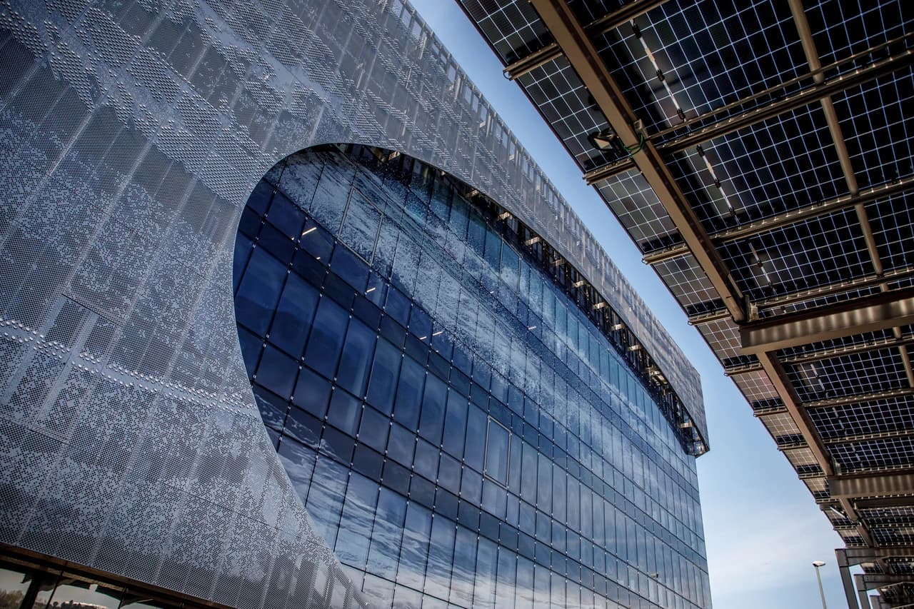 Vue rapprochée d'une façade de bâtiment moderne avec un mur perforé gris argenté et une large section de verre incurvée bleu foncé, reflétant le ciel. Des panneaux solaires sont visibles sur une structure métallique à droite, sous un ciel bleu clair.