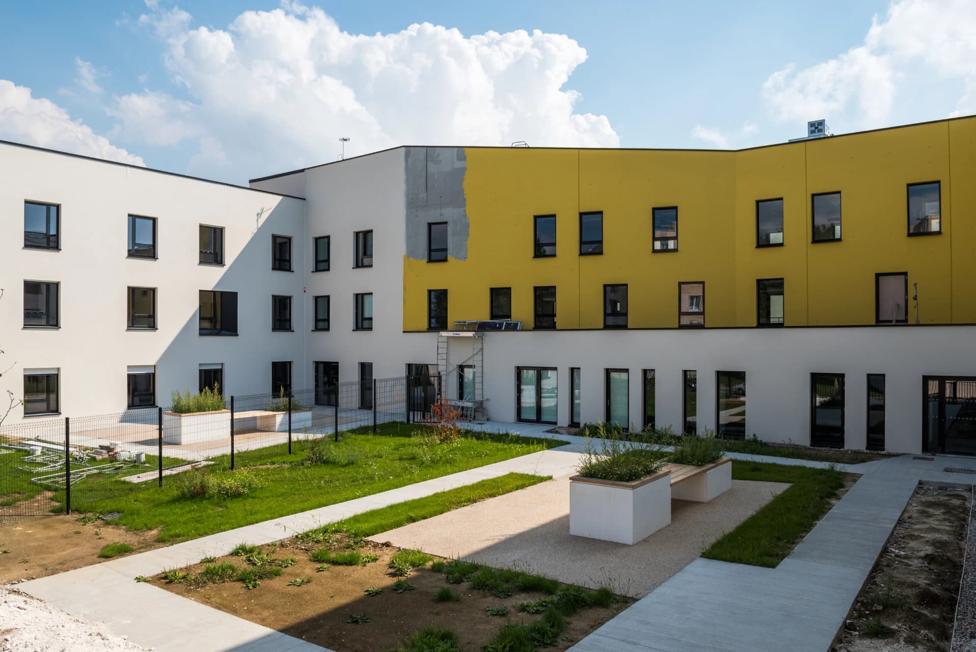 Façade extérieure d'un bâtiment moderne en forme de L, avec une section supérieure jaune vif, des murs blancs et une zone de béton brut apparente, entourant une cour aménagée. Un échafaudage est positionné contre la façade jaune sous un ciel bleu nuageux.