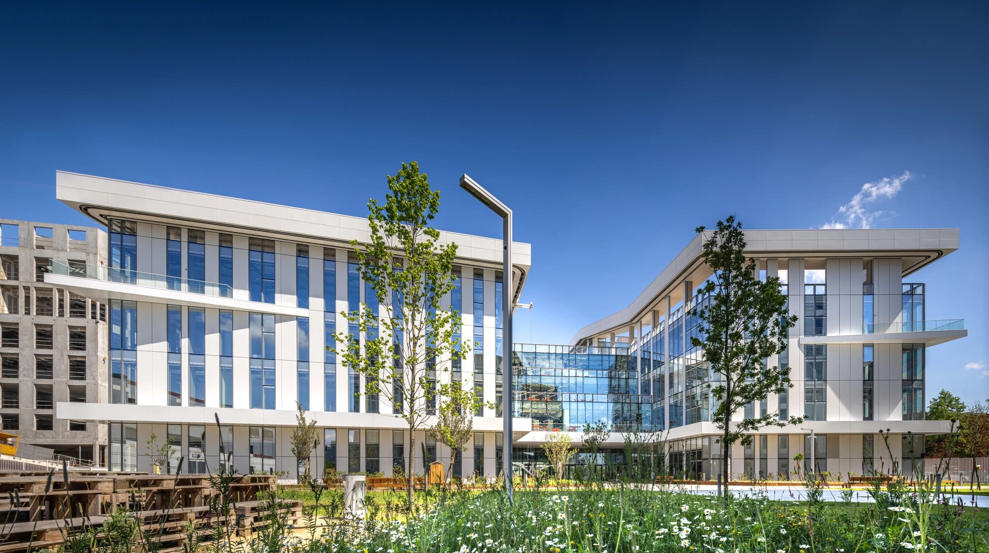 Vue panoramique d'un complexe de bâtiments modernes aux façades blanches et vitrées, avec des lignes incurvées, encadré par des pelouses fleuries et des arbres sous un ciel bleu éclatant.
