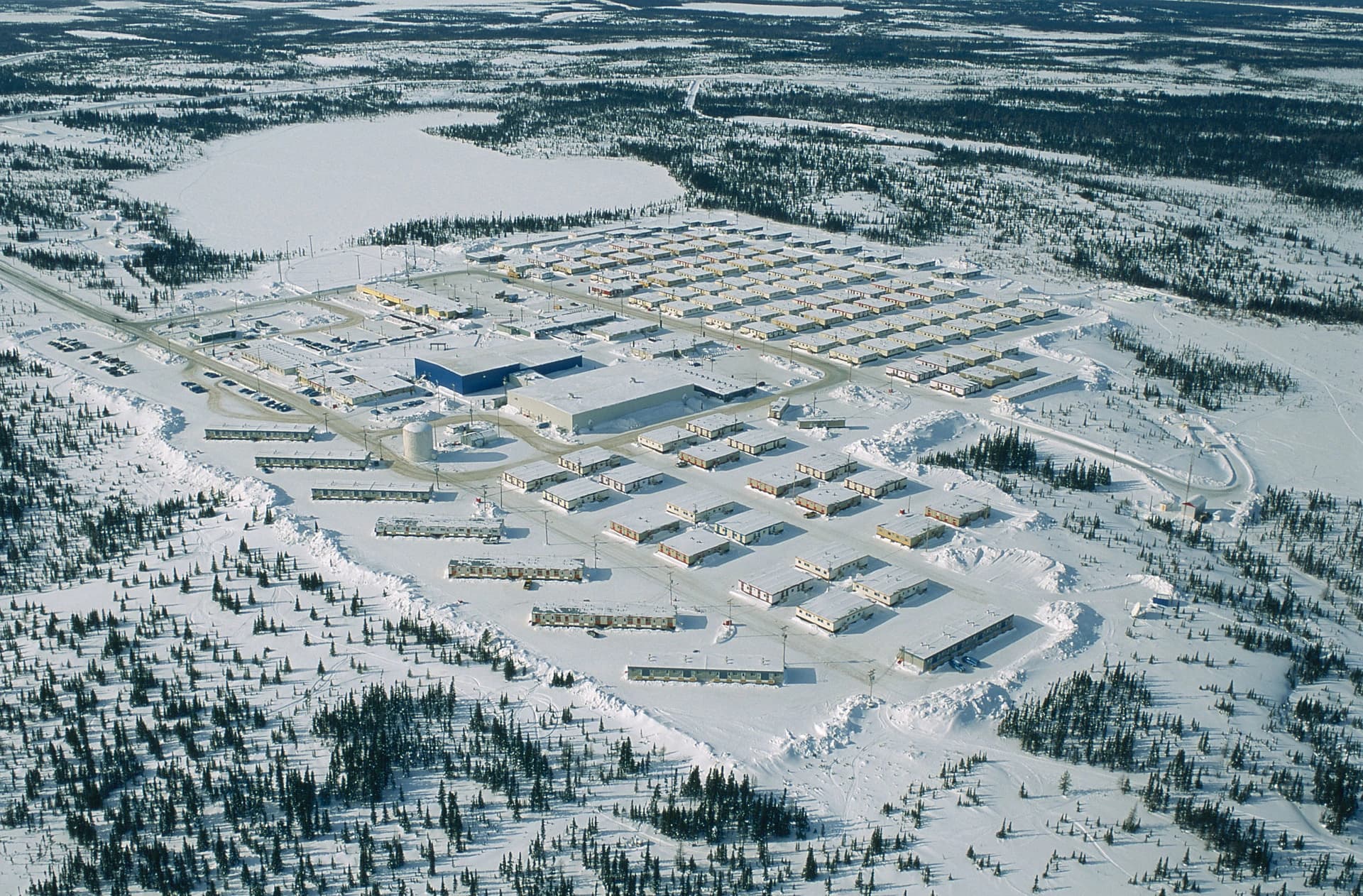 Vue aérienne d'un grand campement ou d'une petite ville d'hiver, comprenant de nombreuses habitations modulaires claires et des bâtiments industriels (dont un bleu vif), s'étendant sur un paysage enneigé bordé de forêts denses et d'un lac gelé.