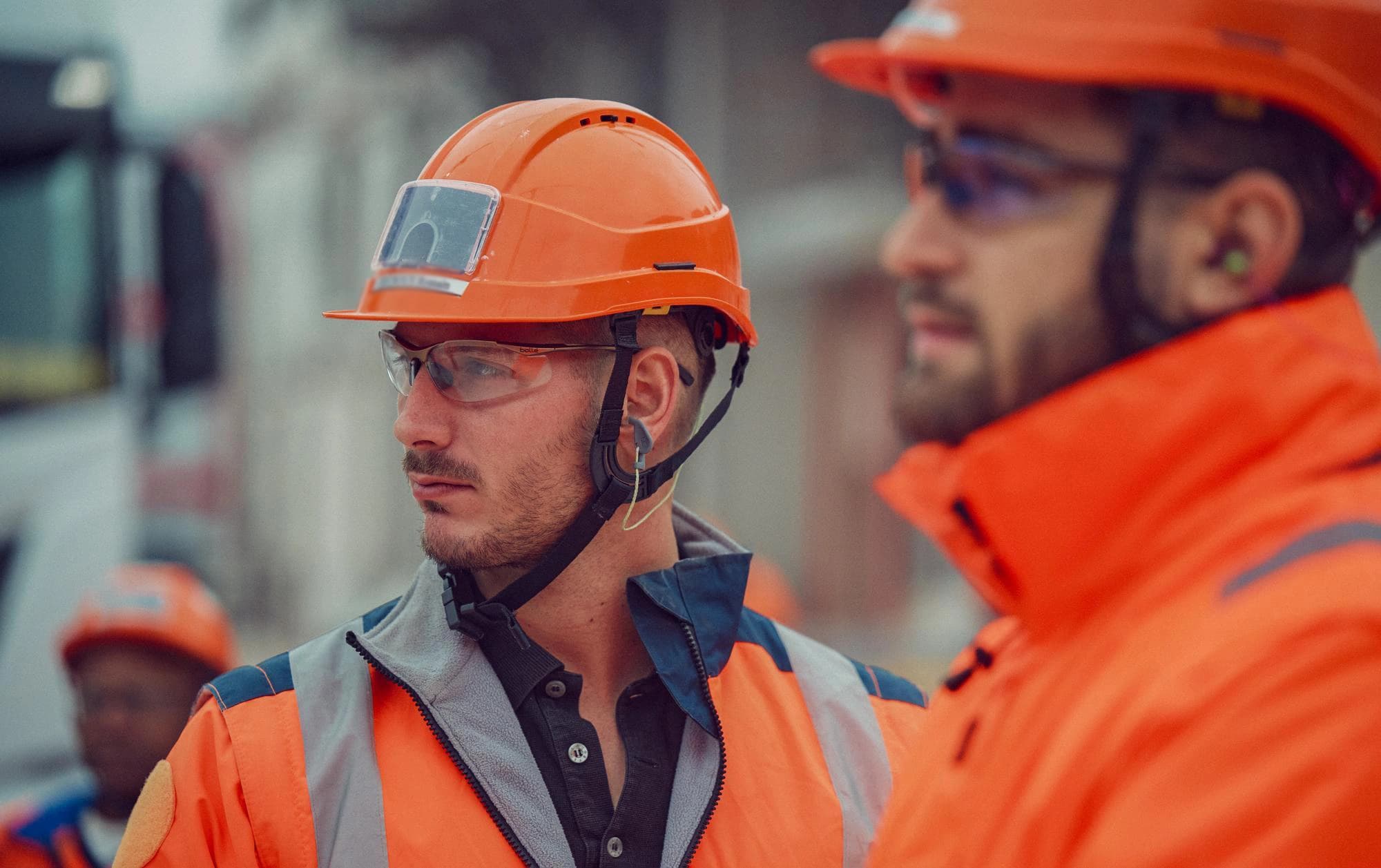 Un homme en tenue de chantier orange, avec un casque, des lunettes de sécurité et des bouchons d'oreille, regarde attentivement à gauche, un collègue flou également en tenue orange étant visible en arrière-plan.