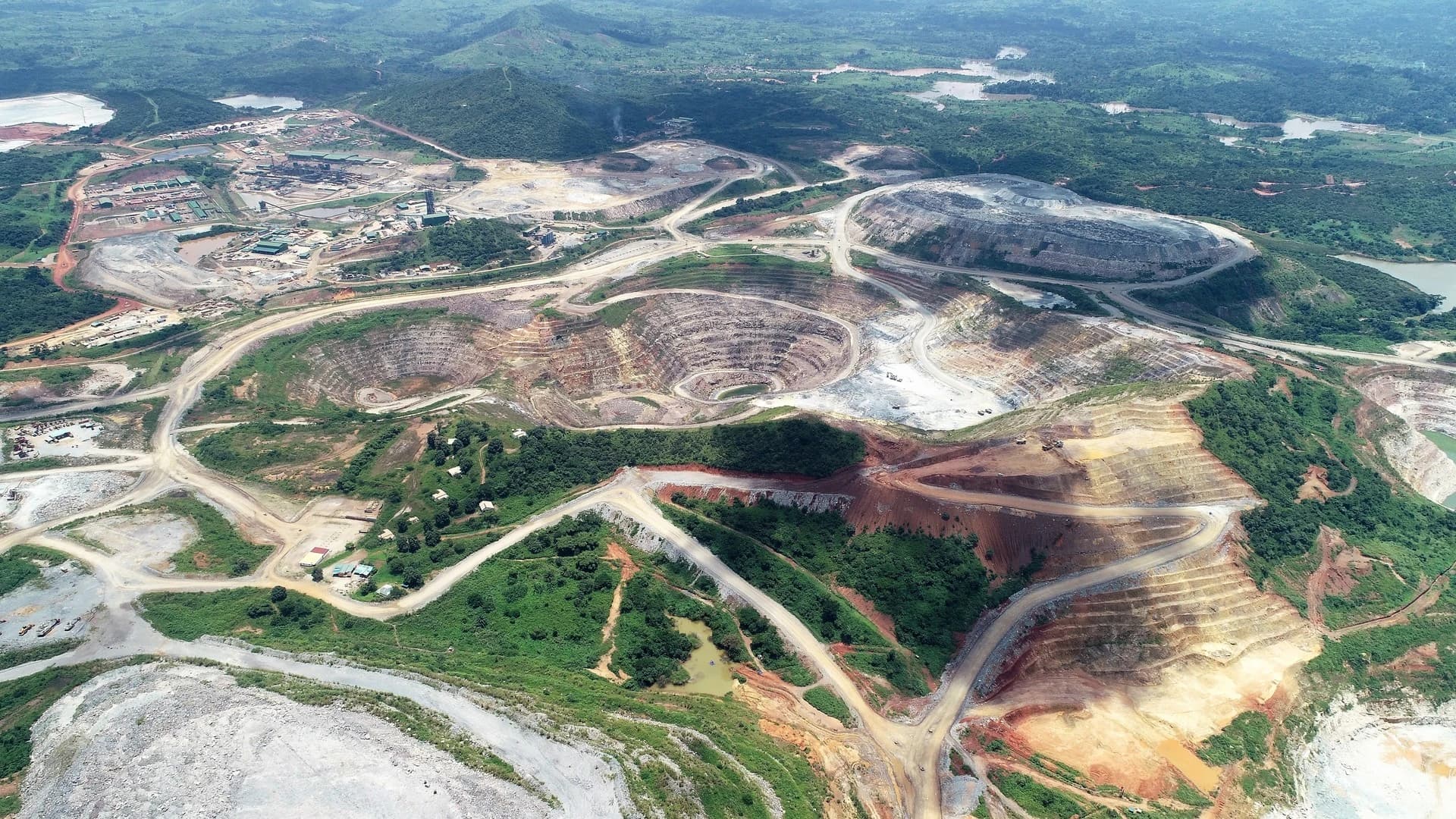 Vue aérienne d'une immense mine à ciel ouvert, présentant de multiples fosses terrassées aux couleurs variées (rouge, ocre, gris) traversées par des routes, au milieu de paysages verdoyants et de plans d'eau.
