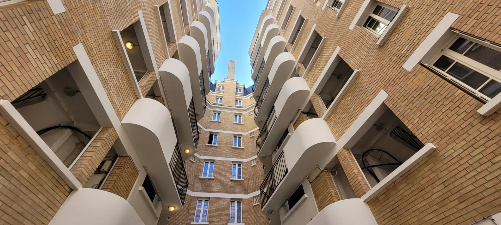 Vue en contre-plongée d'une cour intérieure montrant des façades en briques claires avec des balcons arrondis blancs et des balustrades noires, convergeant vers un immeuble central coiffé d'un toit mansardé sous un ciel bleu.