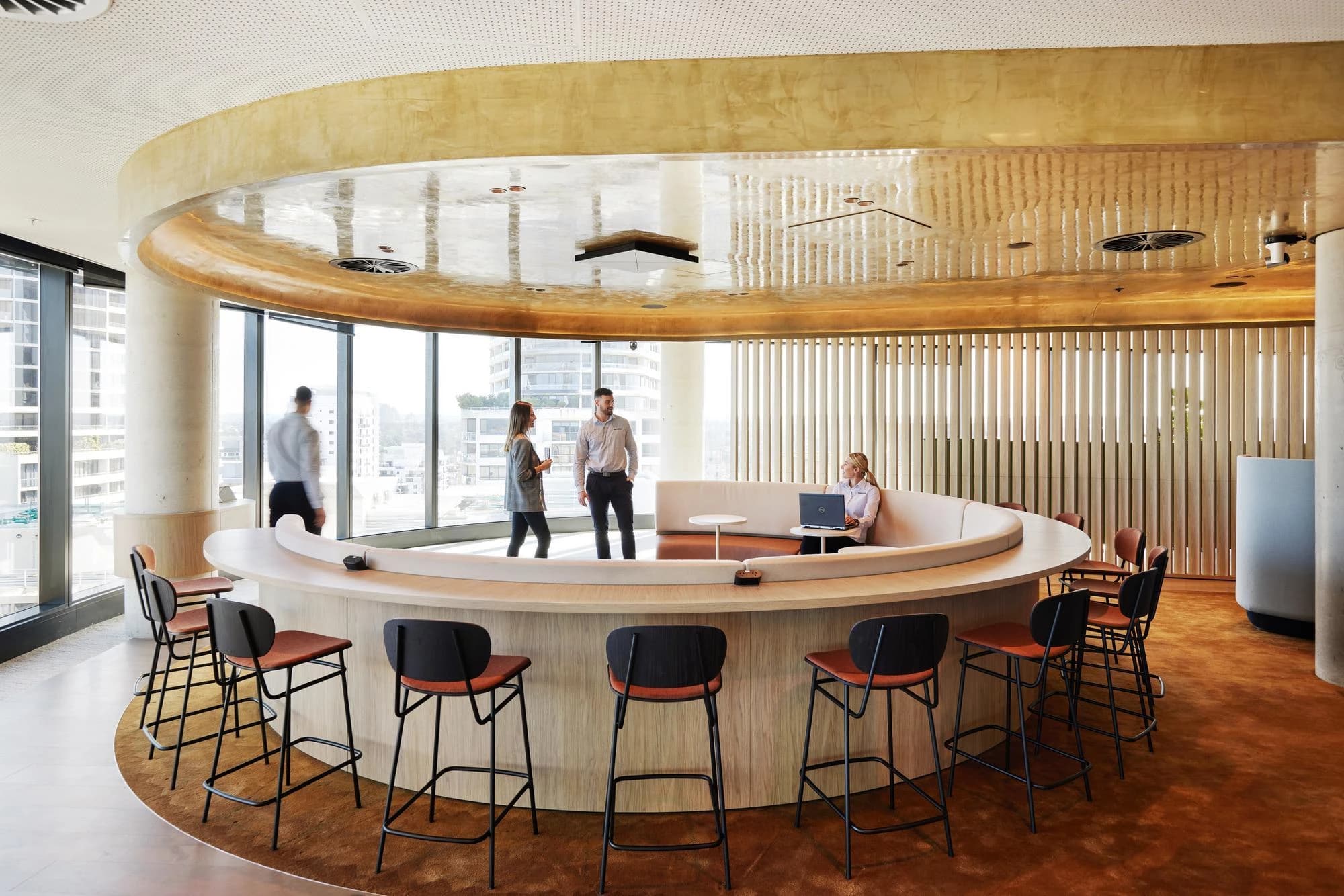 Un salon de bureau moderne et incurvé présente un comptoir en bois clair avec des tabourets marron-orange, un canapé beige et de grandes fenêtres donnant sur une ville. Des personnes travaillent sur des ordinateurs portables ou conversent dans cet espace aux tons chauds, sous un plafond doré et réfléchissant.